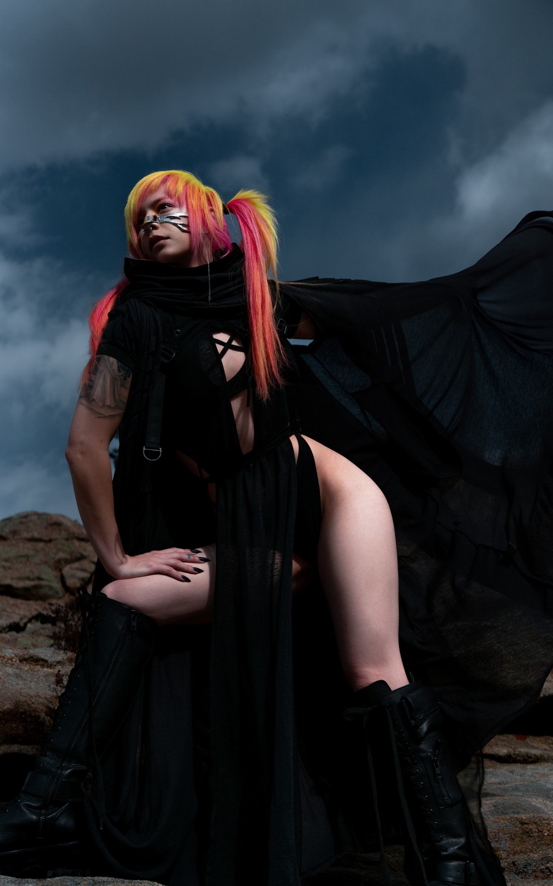 Person with colorful hair and makeup, wearing black clothing and boots, standing outdoors against a cloudy sky.