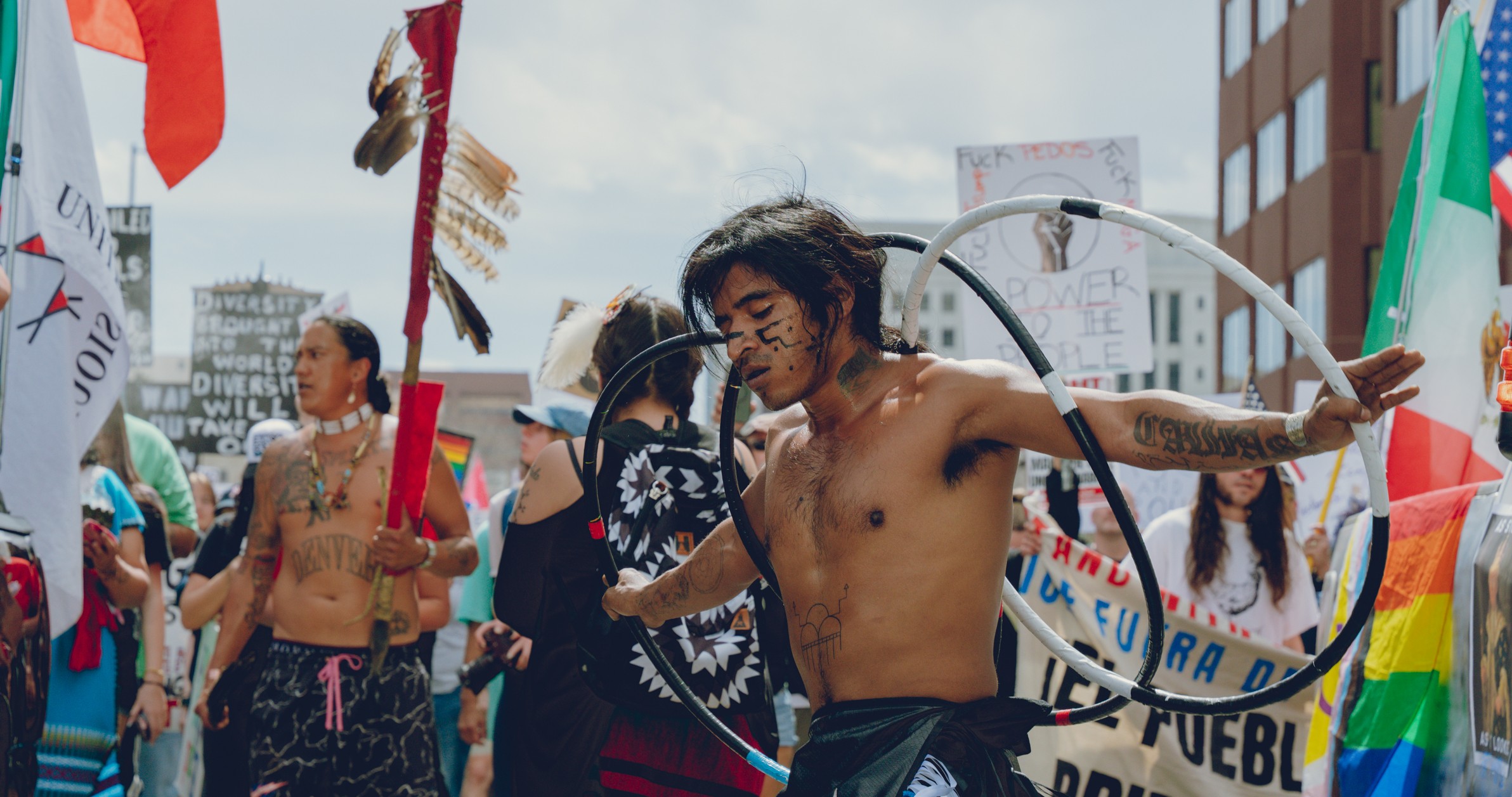 Person with long hair and tattoos hula hooping at a parade or protest, surrounded by people and flags.