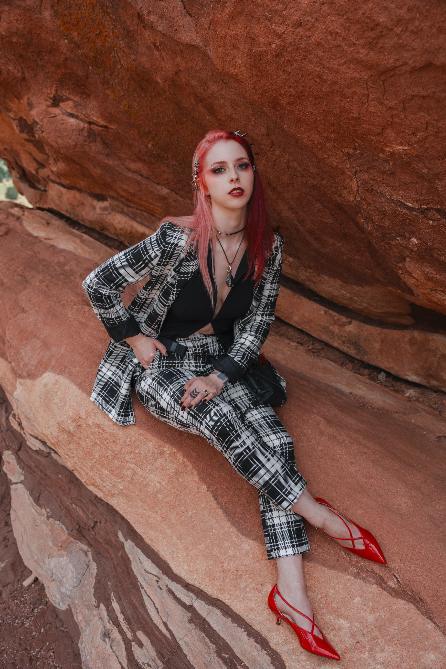 Woman with pink hair in plaid outfit sitting on rock, red shoes, desert background.