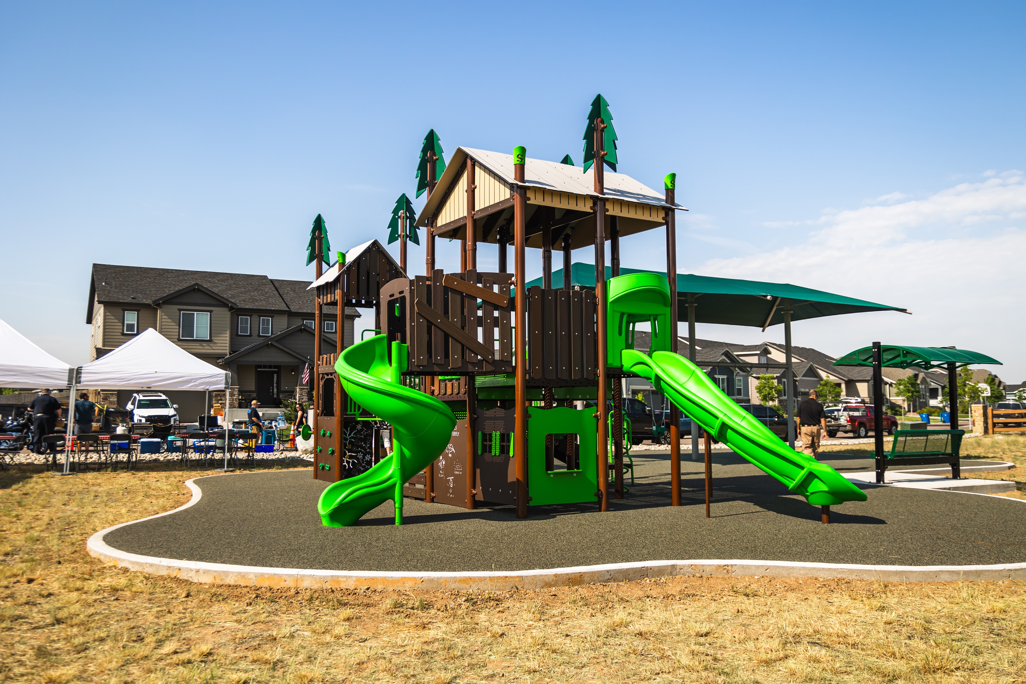 Colorful playground structure with slides and climbing areas, surrounded by a safety surface and residential buildings in the background.