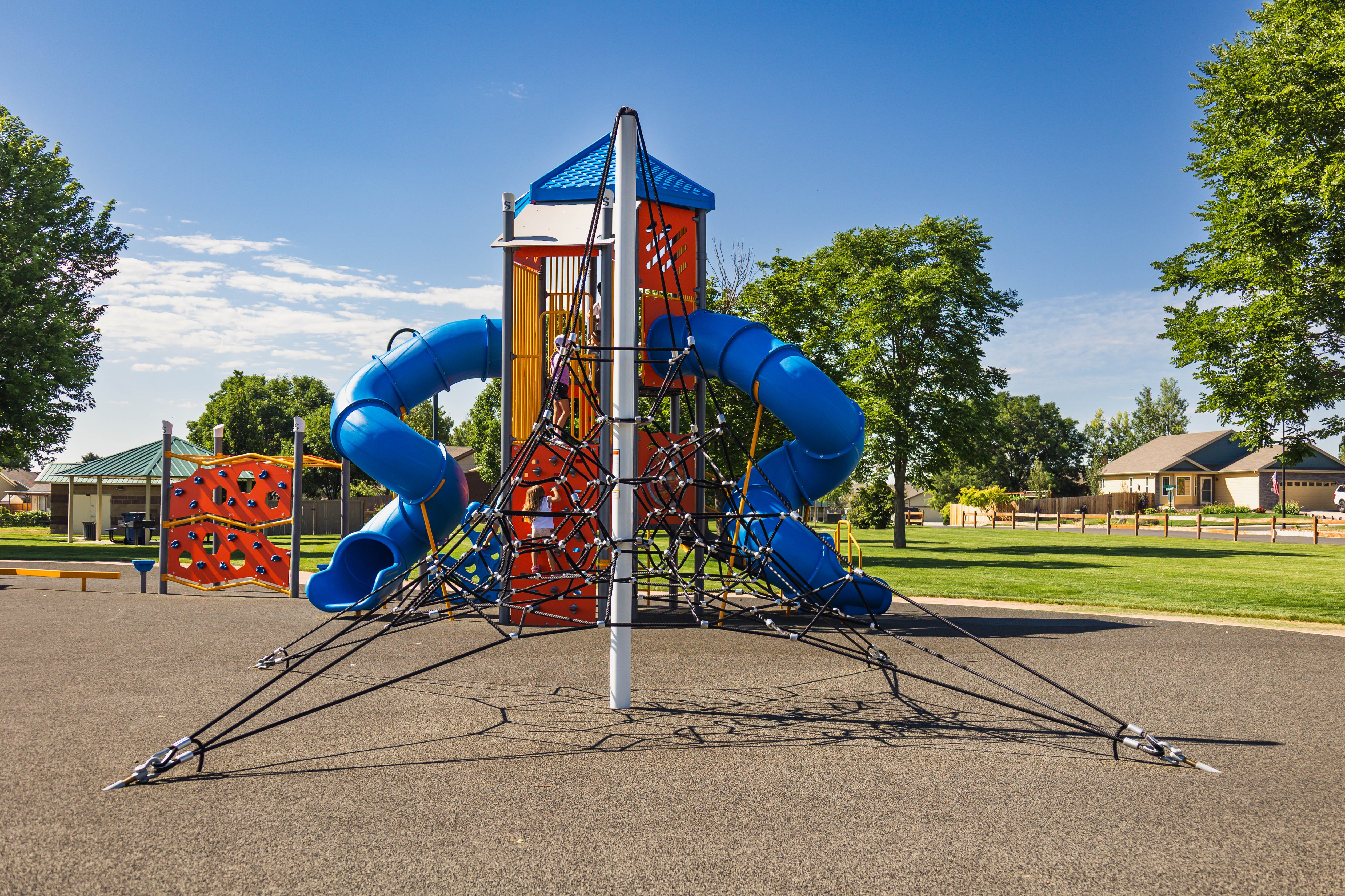 Colorful playground structure with multiple blue slides, climbing areas, and a tower, set in a park with trees and houses nearby.