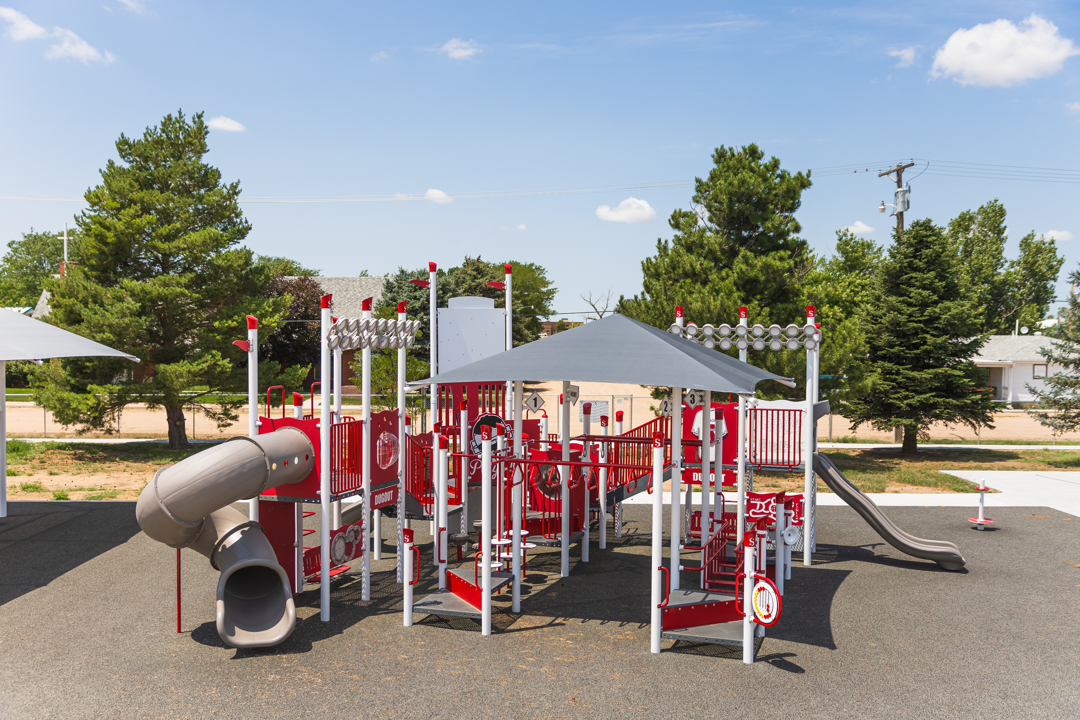 Colorful playground with slides, climbing structures, and shade, set in an outdoor area with trees and a clear sky.