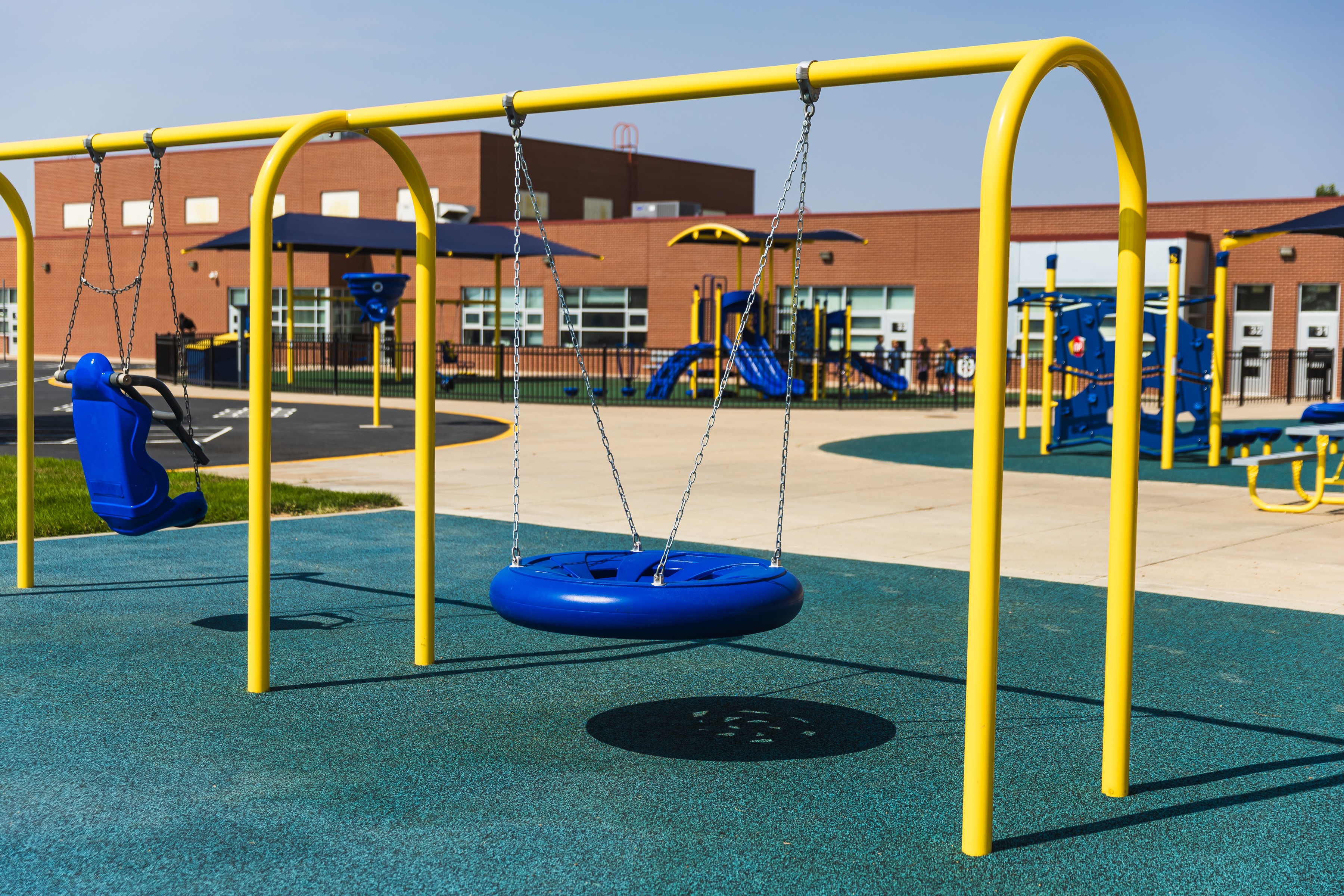 Empty playground with yellow and blue swings, slides, and exercise equipment, with a school building in the background.