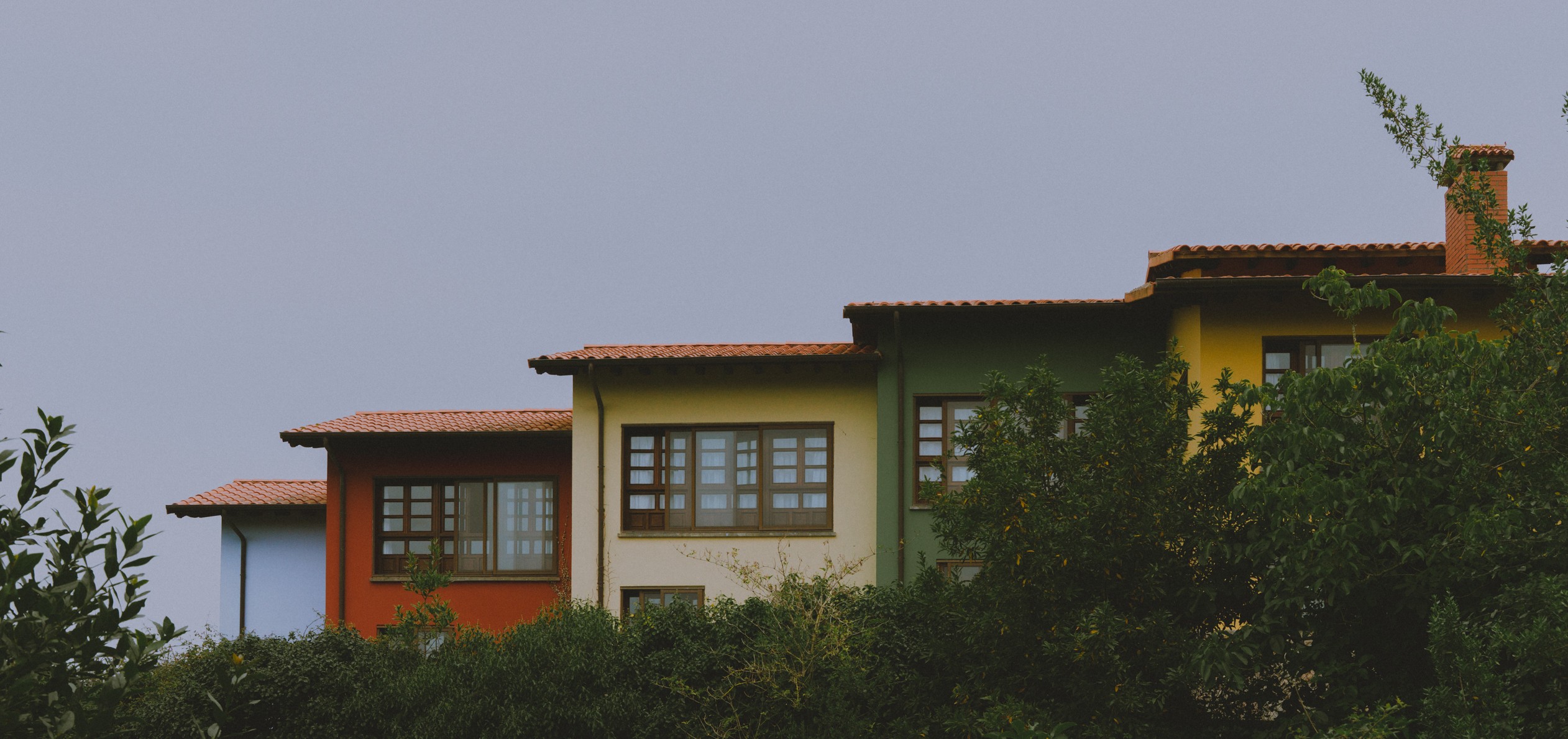 Three colorful houses with trees in front, under a cloudy sky.