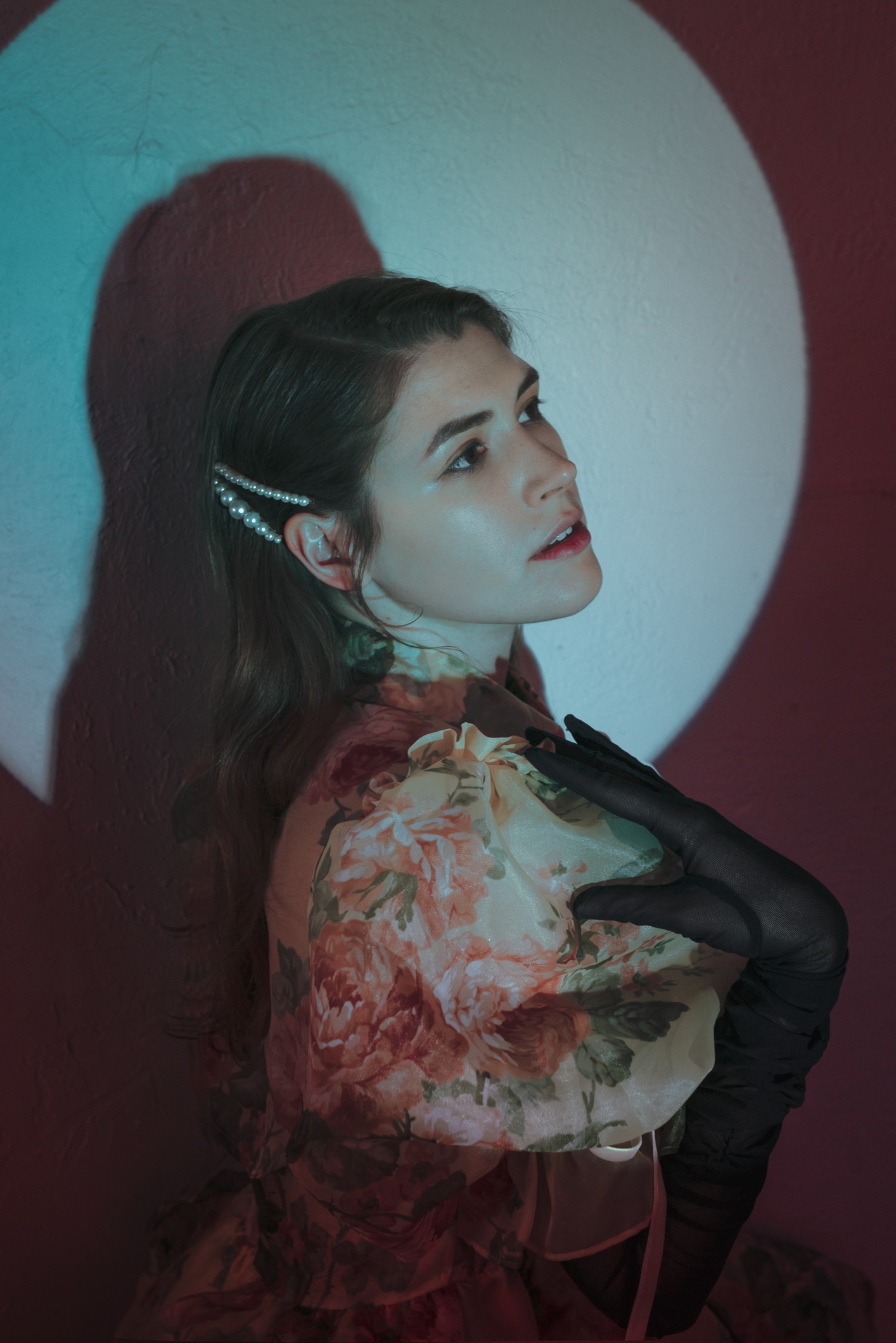 Young woman with dark hair, pearl hair accessory, wearing floral dress and black gloves, poses against a colorful background.