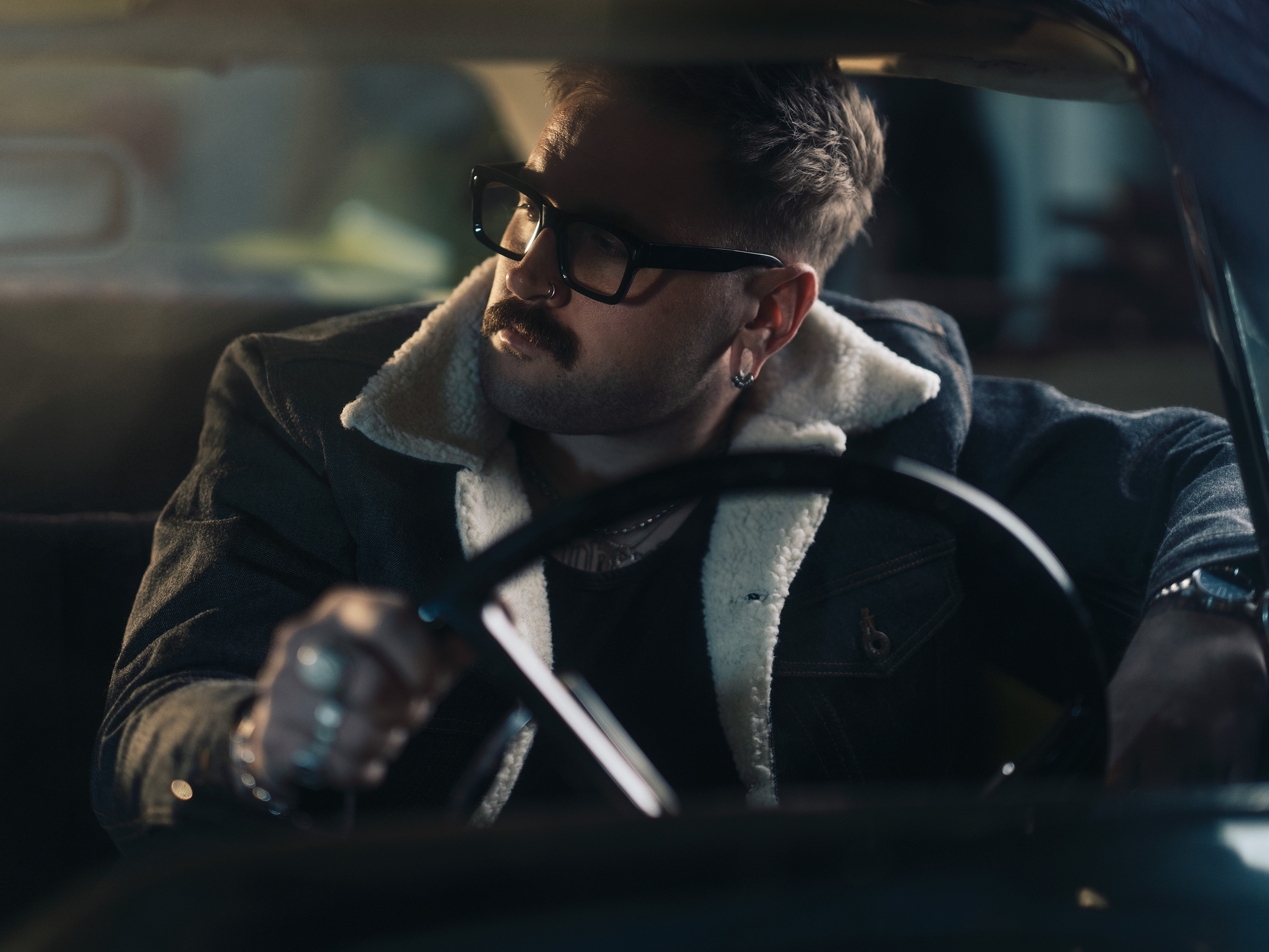 Man with glasses and a beard driving a car, wearing a jacket with a fur collar, looking to the side.