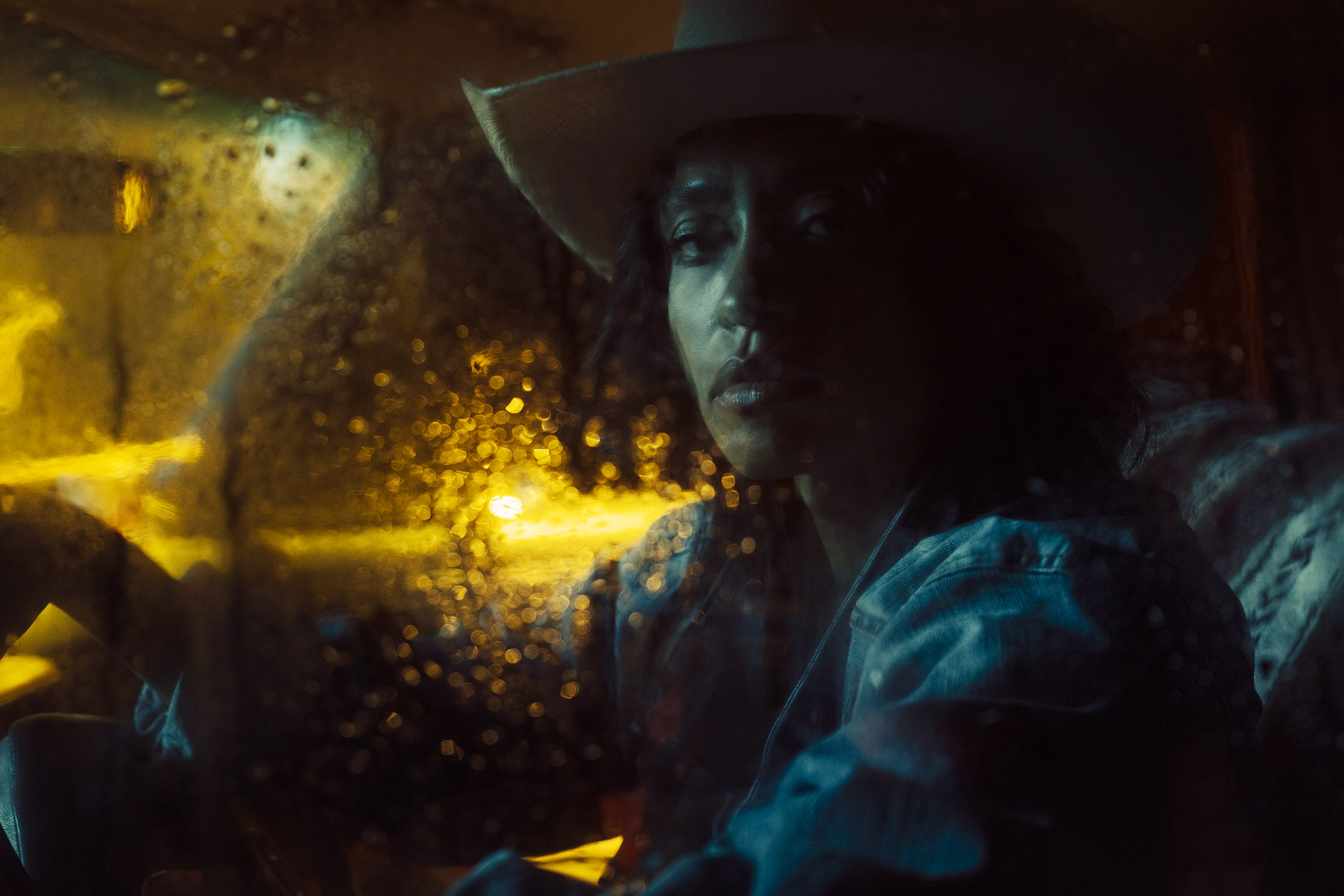 Woman wearing a wide-brimmed hat sitting in a vehicle at night, rain on window with yellow streetlights outside.