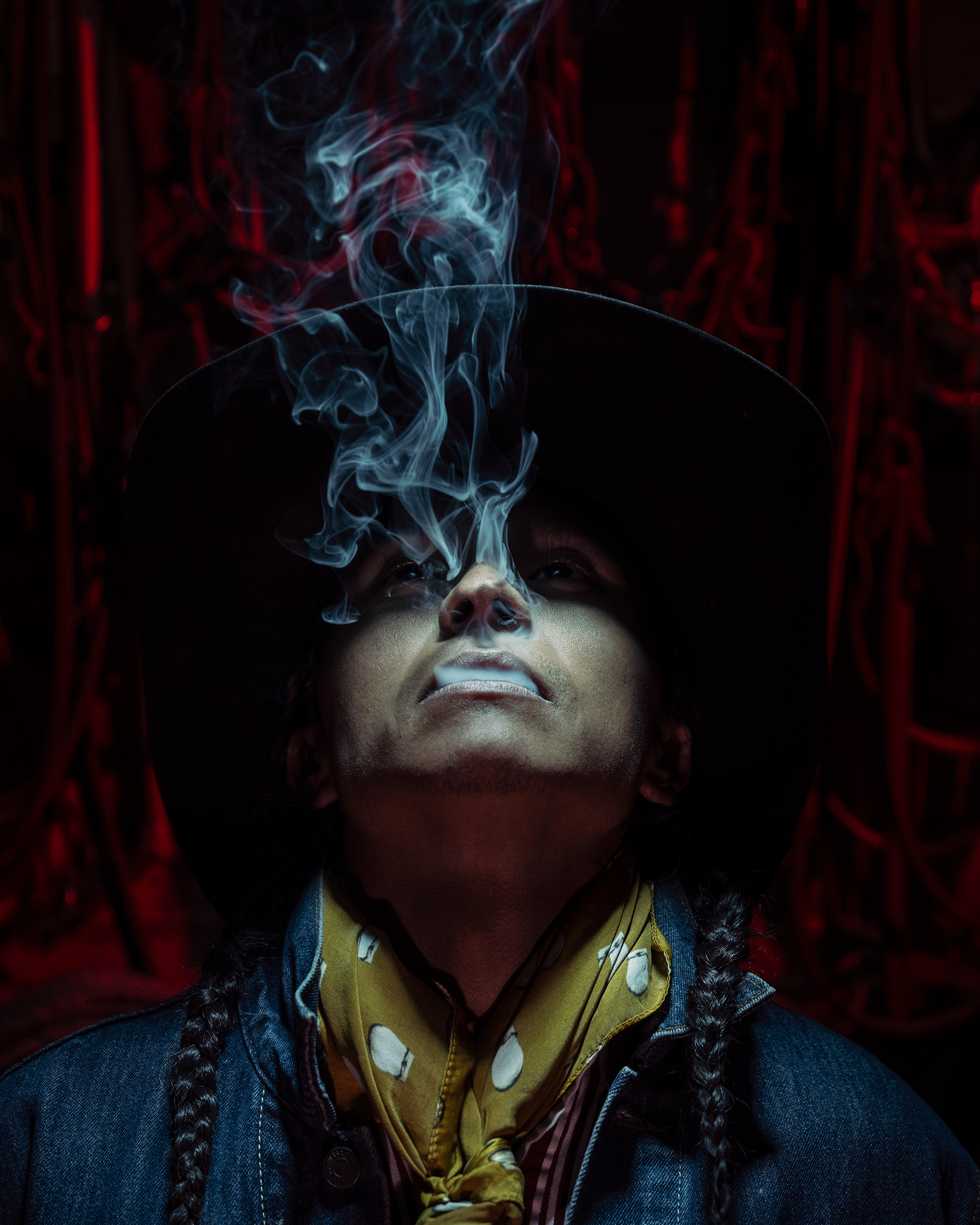 Person wearing a wide-brimmed hat and yellow bandana, smoking with smoke rising, dark background with red elements.