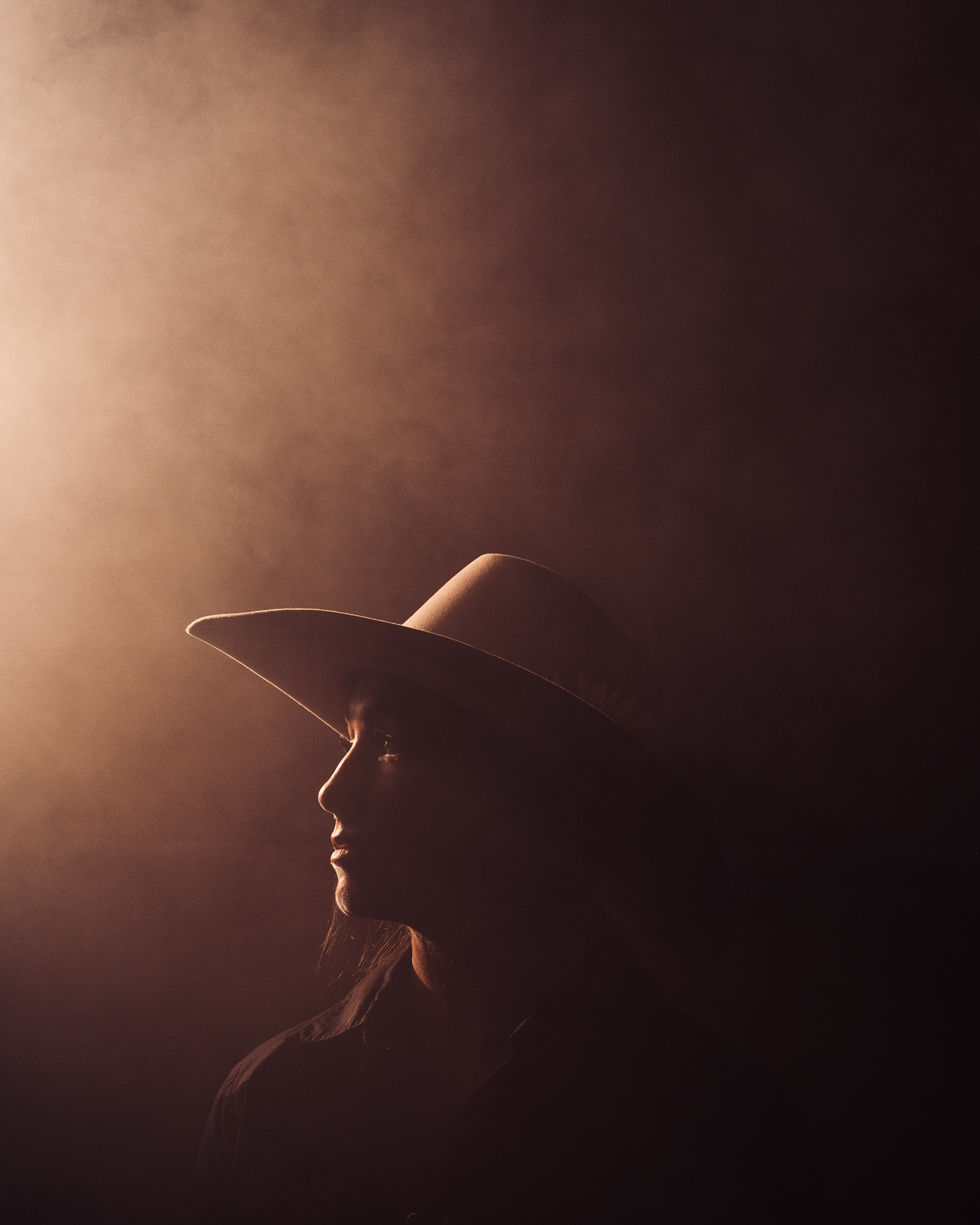 Silhouette of a person wearing a wide-brimmed hat, facing left, with a dark background and soft lighting.