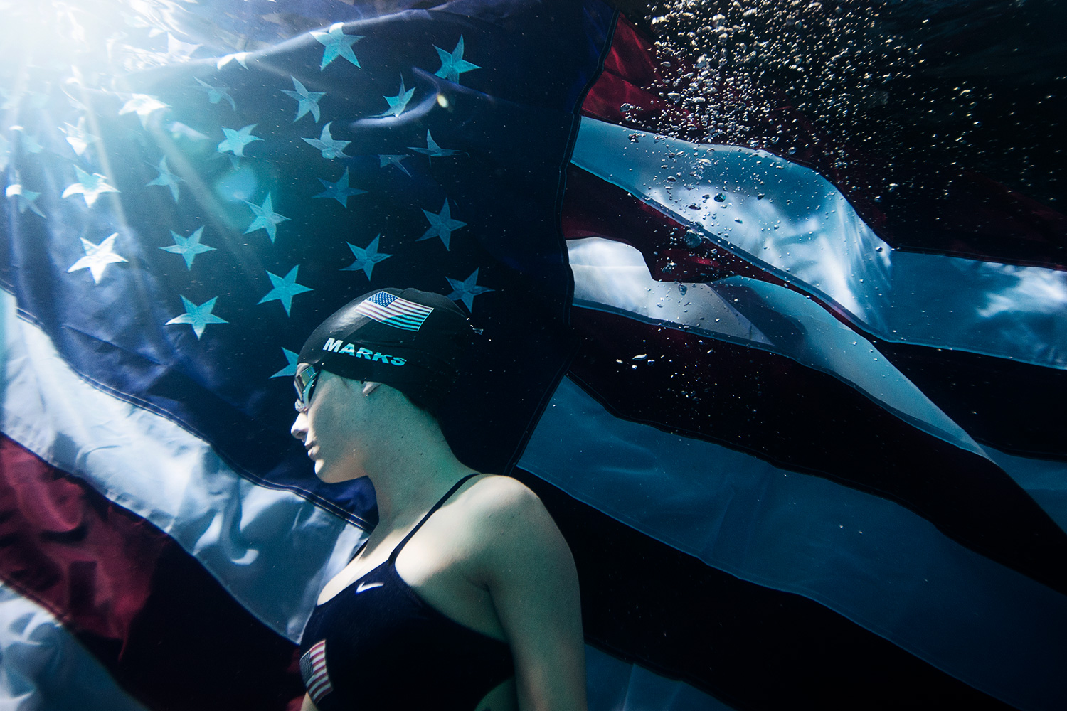Person lying on an American flag with water droplets, wearing a swim cap and goggles, in a pool or water setting.