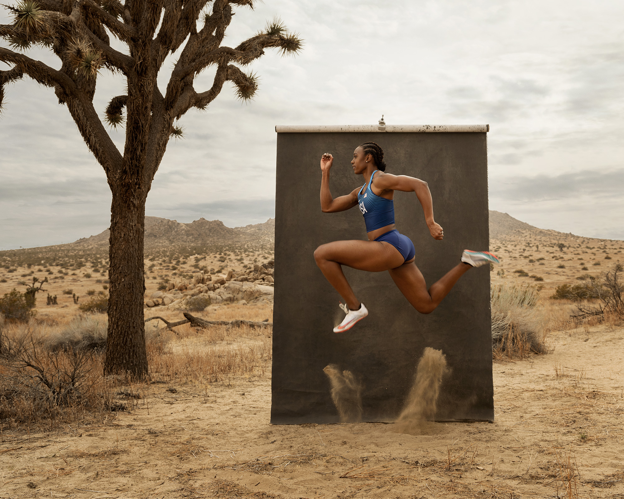 A woman in athletic clothing jumps in front of a black backdrop in a desert landscape with a tree and mountains.