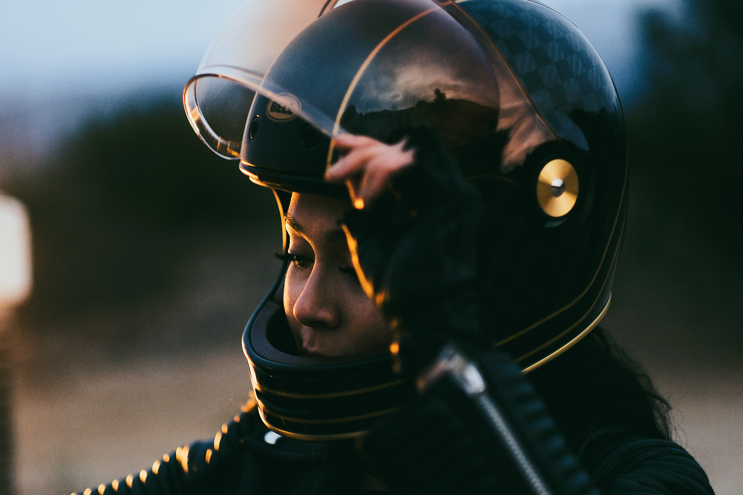 A person wearing a black motorcycle helmet with a clear visor, outdoors during sunset or dusk, with a blurred background.