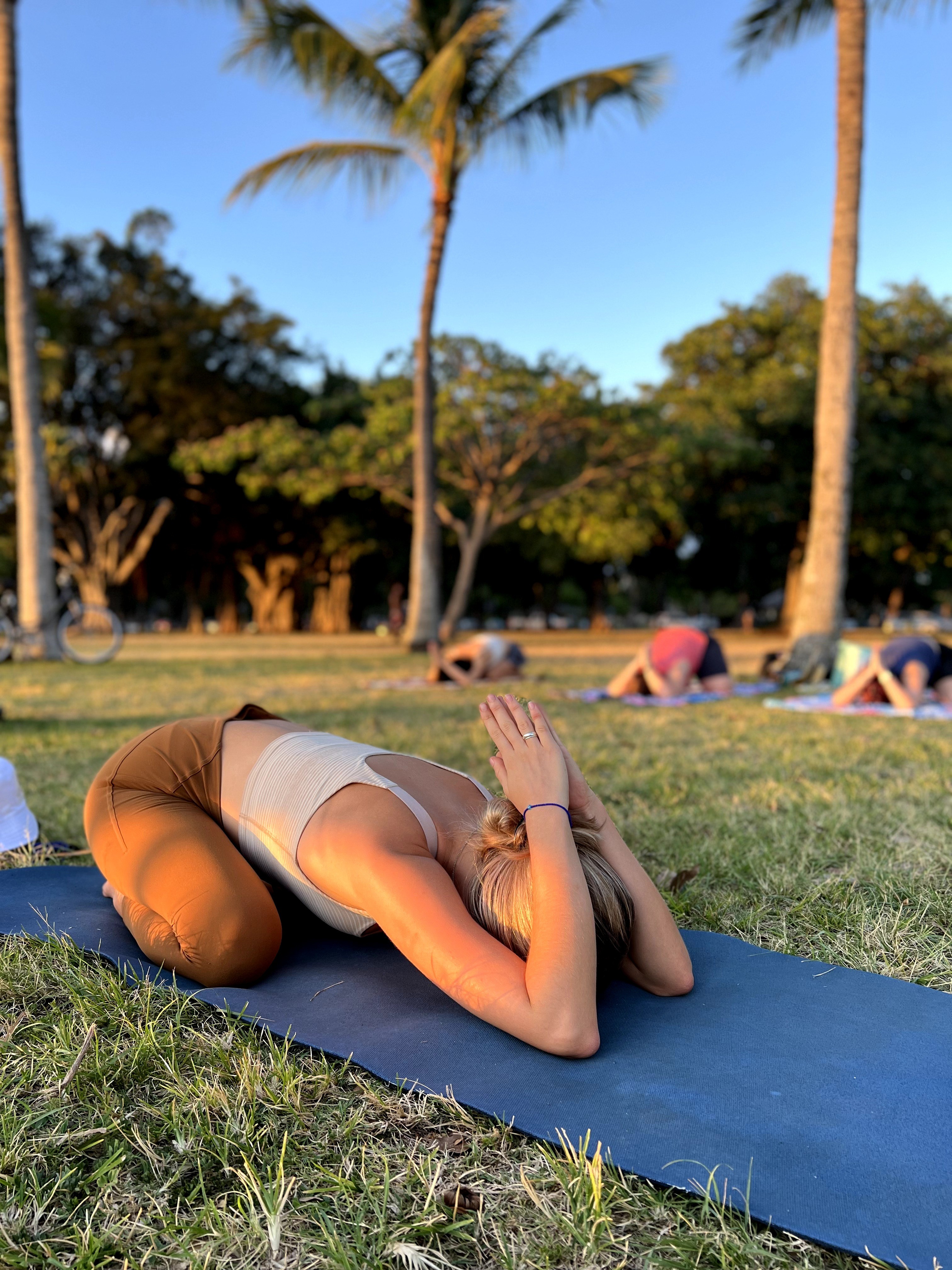 Person practicing yoga outdoors on a mat, surrounded by trees and other people, during daytime.