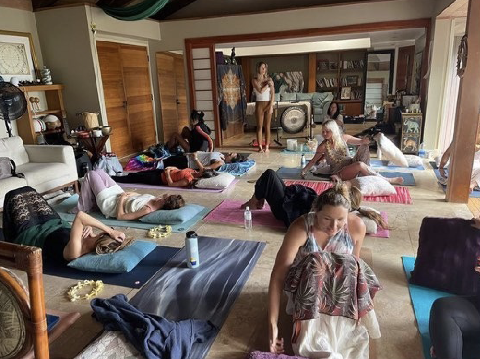 Group of people practicing yoga on mats in a living room with furniture and decorations, some lying down and others sitting or standing.