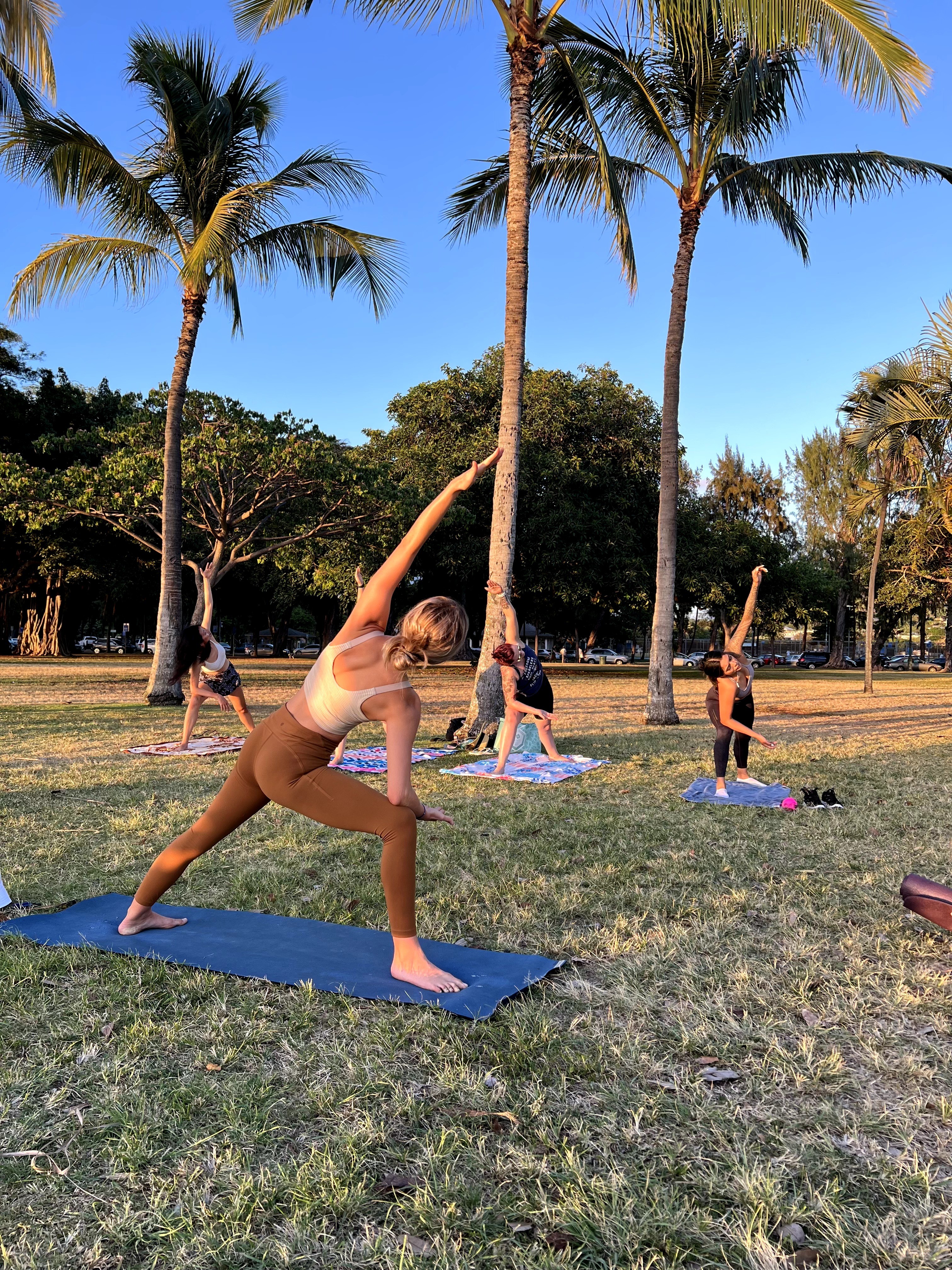 Group of people practicing yoga outdoors on mats in a park with palm trees and blue sky.