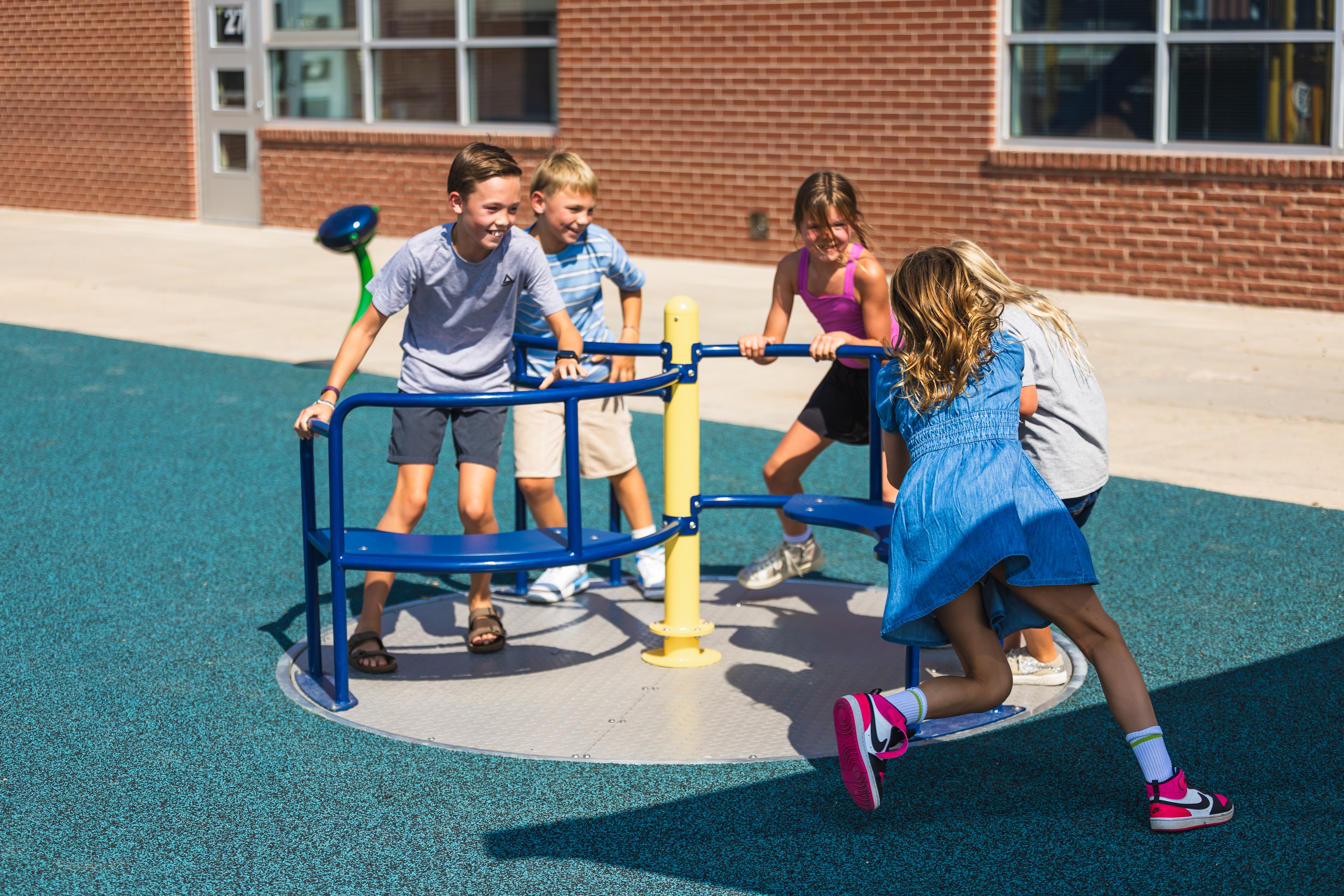 Children playing on a yellow and blue merry-go-round outside a brick building, with a girl in a blue dress running nearby.