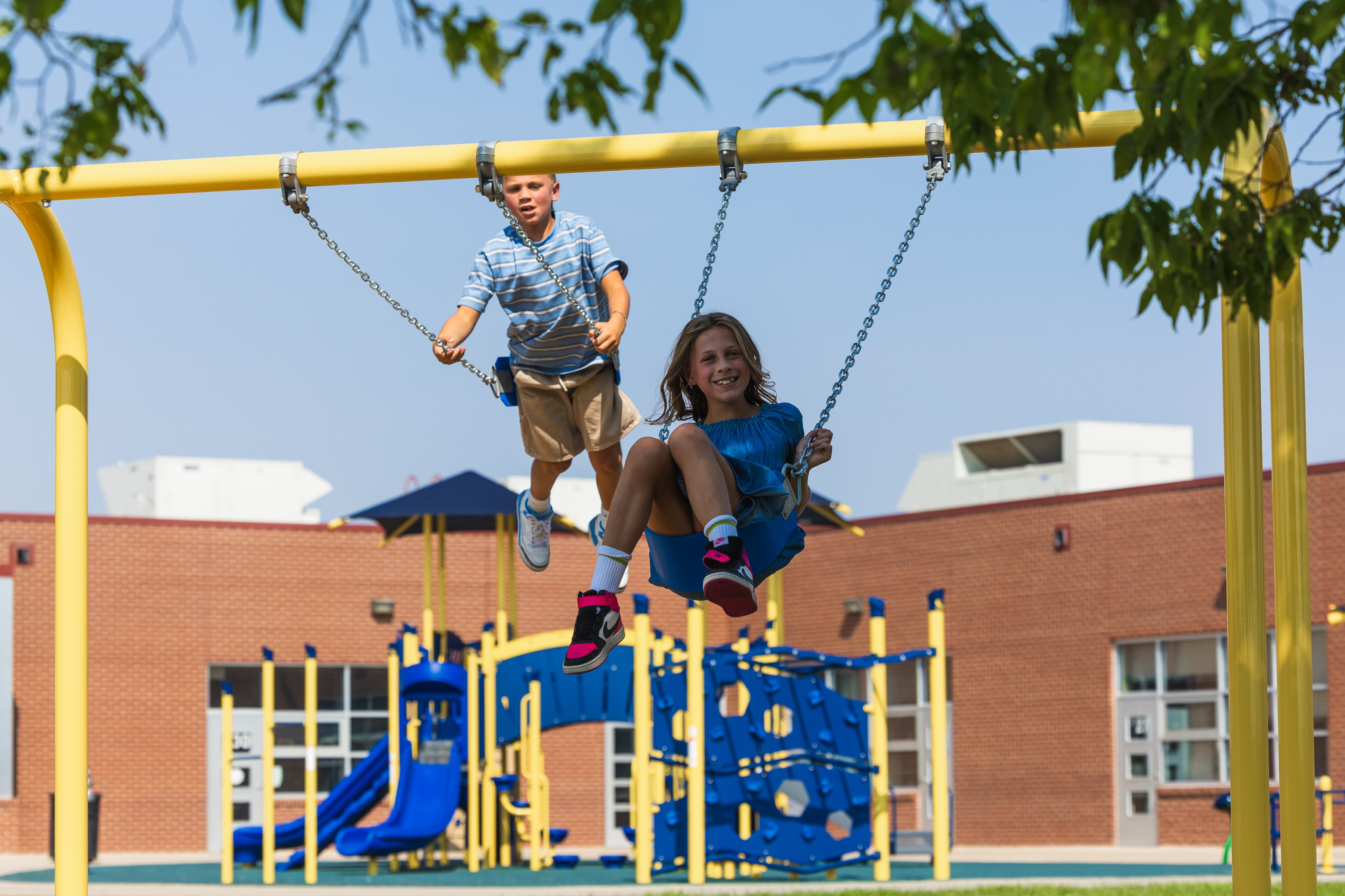 Two children on a swing set at a playground, with a building and trees in the background.