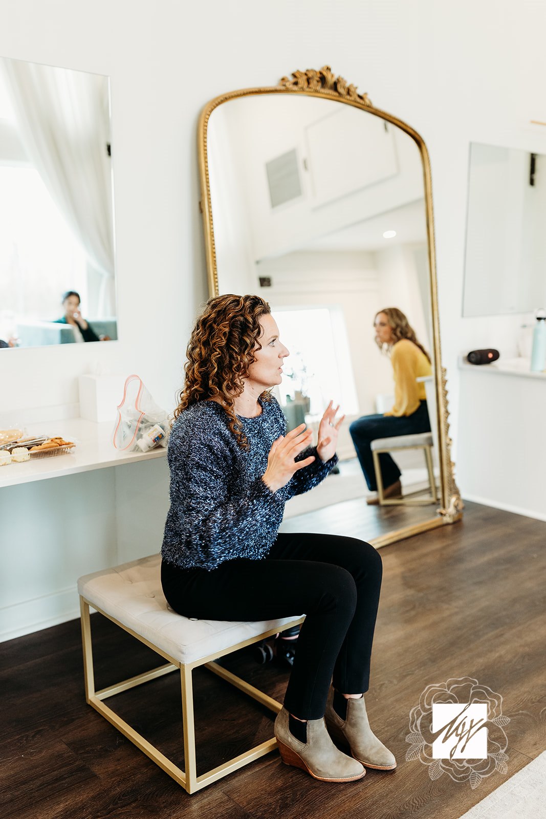 Woman with curly hair sitting on a bench, talking, with a large mirror behind her reflecting her and a yellow chair.