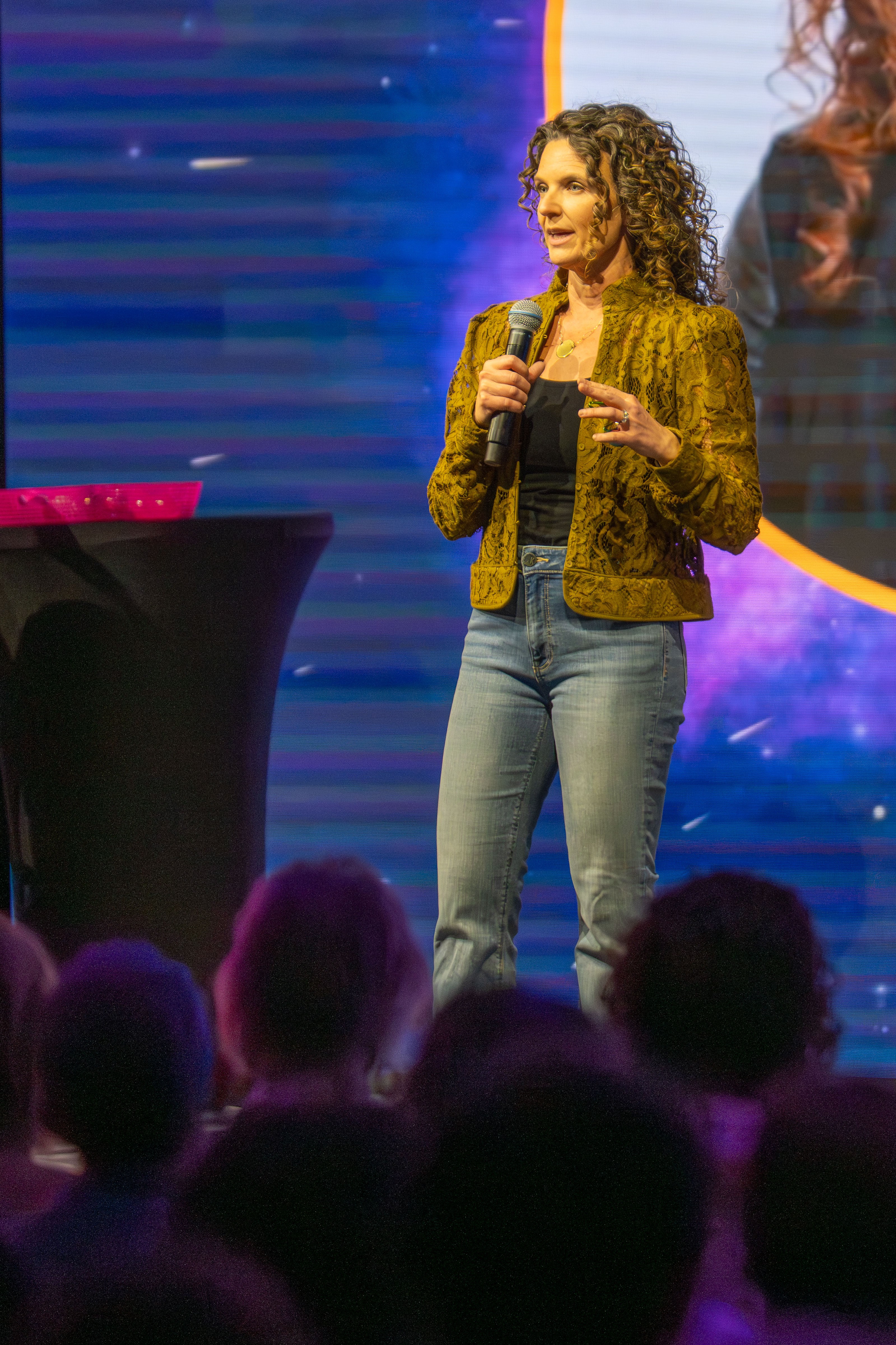 Woman with curly hair speaking into a microphone on stage, audience in foreground, colorful background.