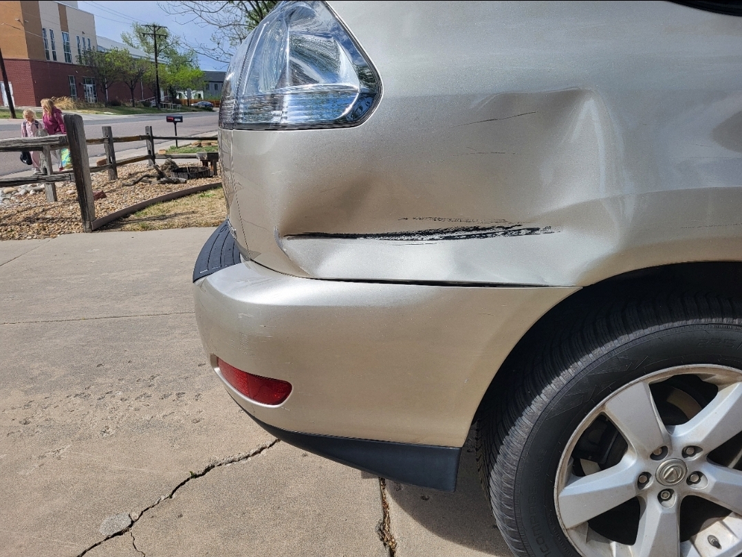 Close-up of a silver car's damaged front right corner with a dented bumper and headlight, parked on a concrete surface.