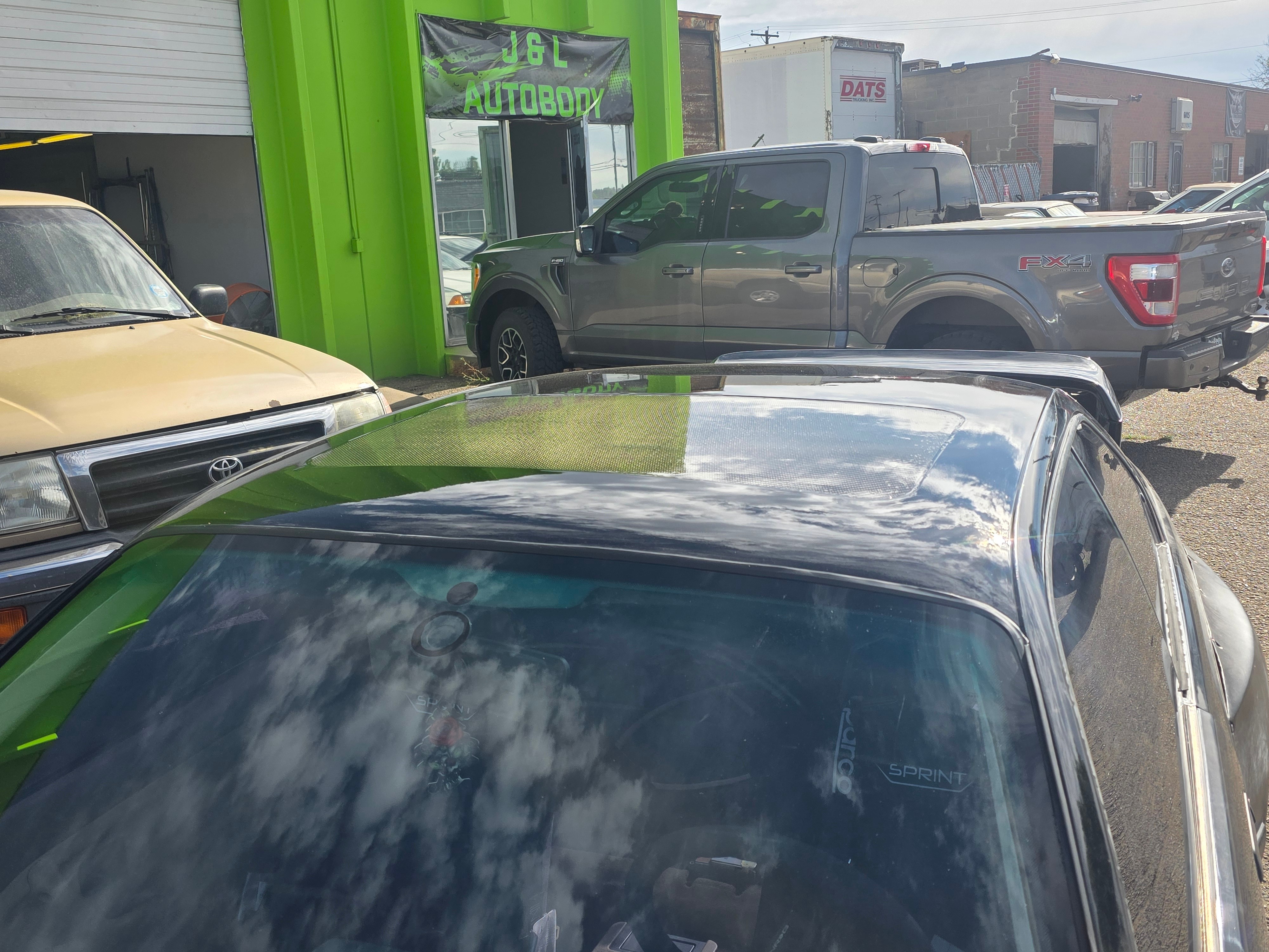 Close-up of a car's windshield reflecting clouds and sky, with other vehicles and a green building in background.