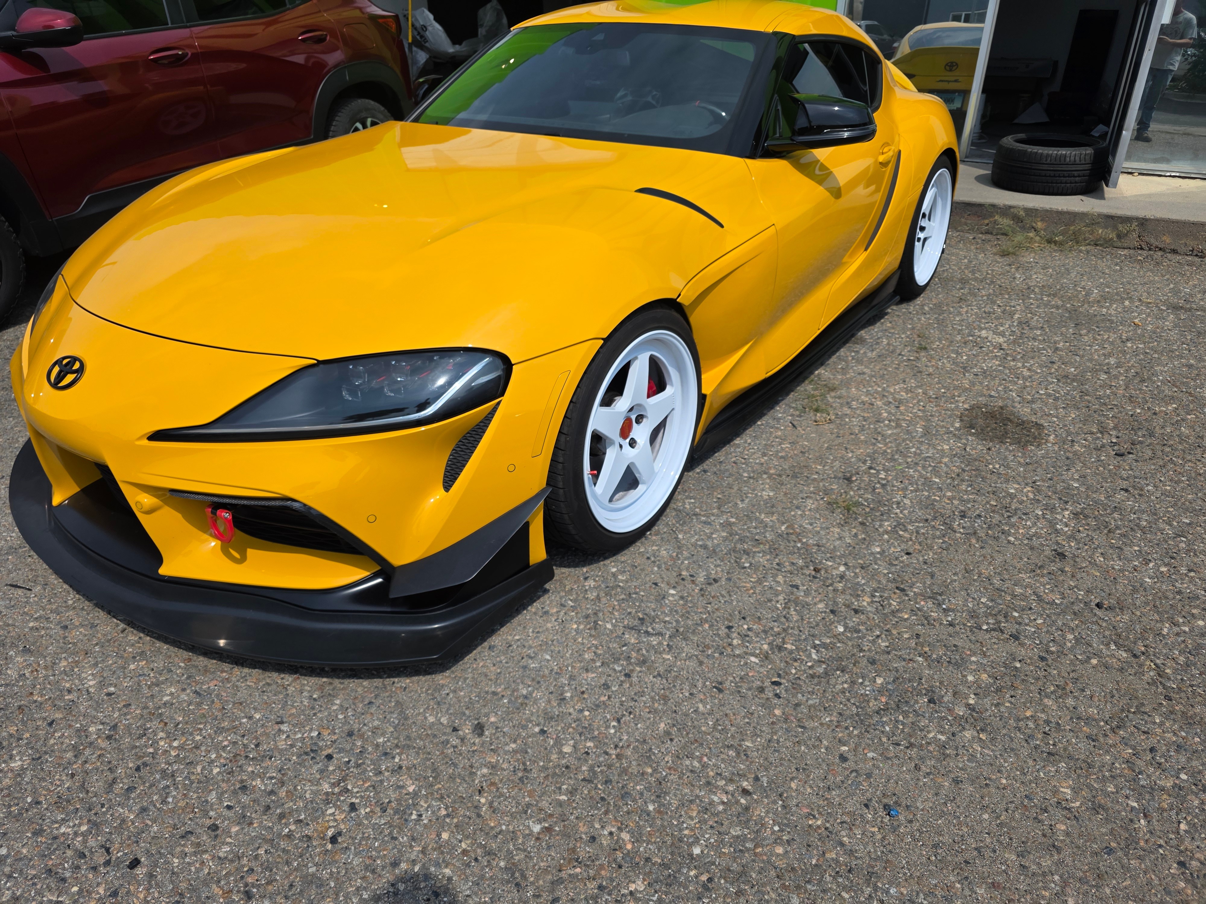 Yellow sports car parked on gravel, with black front splitter and white wheels, in front of a building.