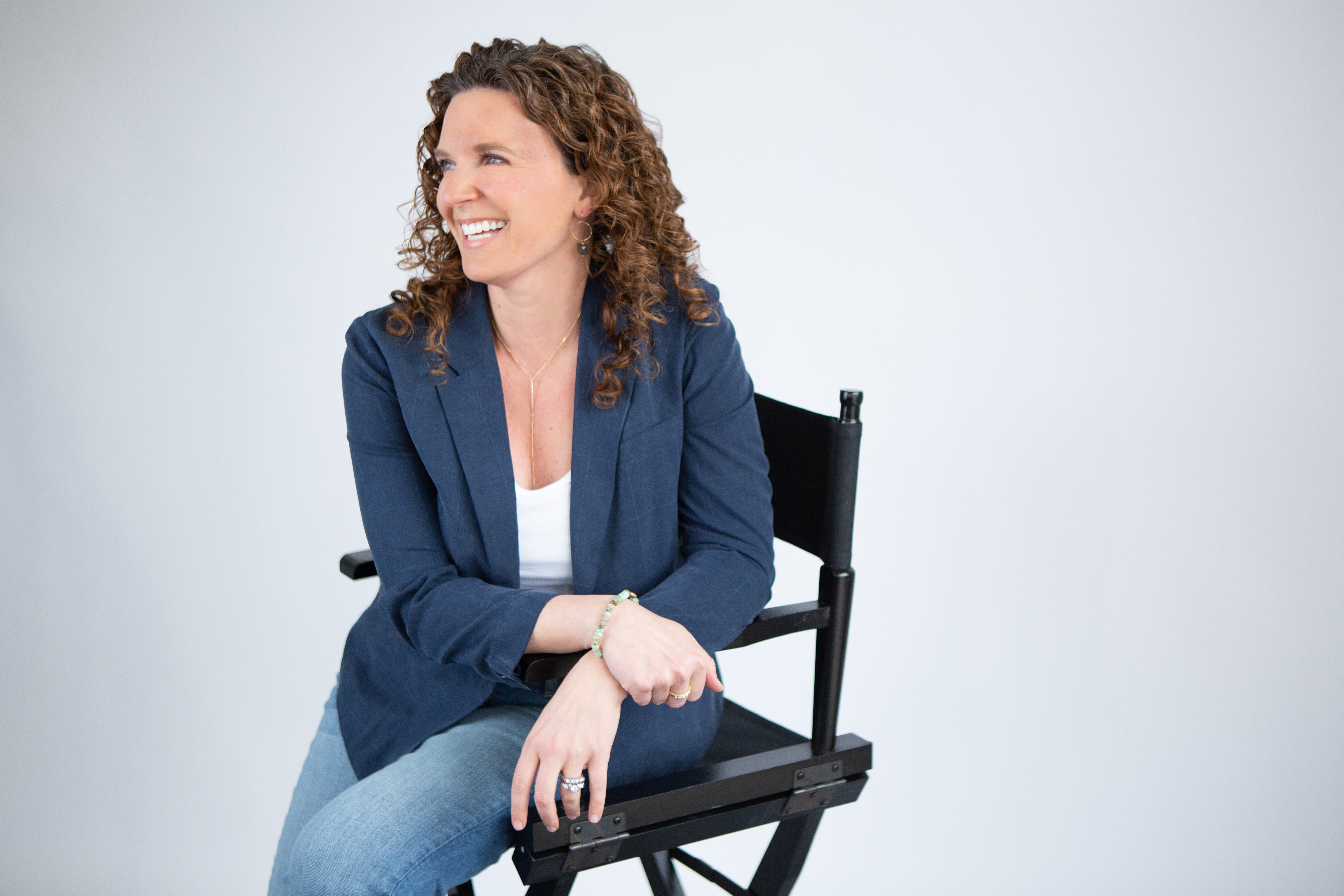 Woman with curly hair smiling, sitting on a black chair, wearing a blue blazer and light jeans, against a plain white background.