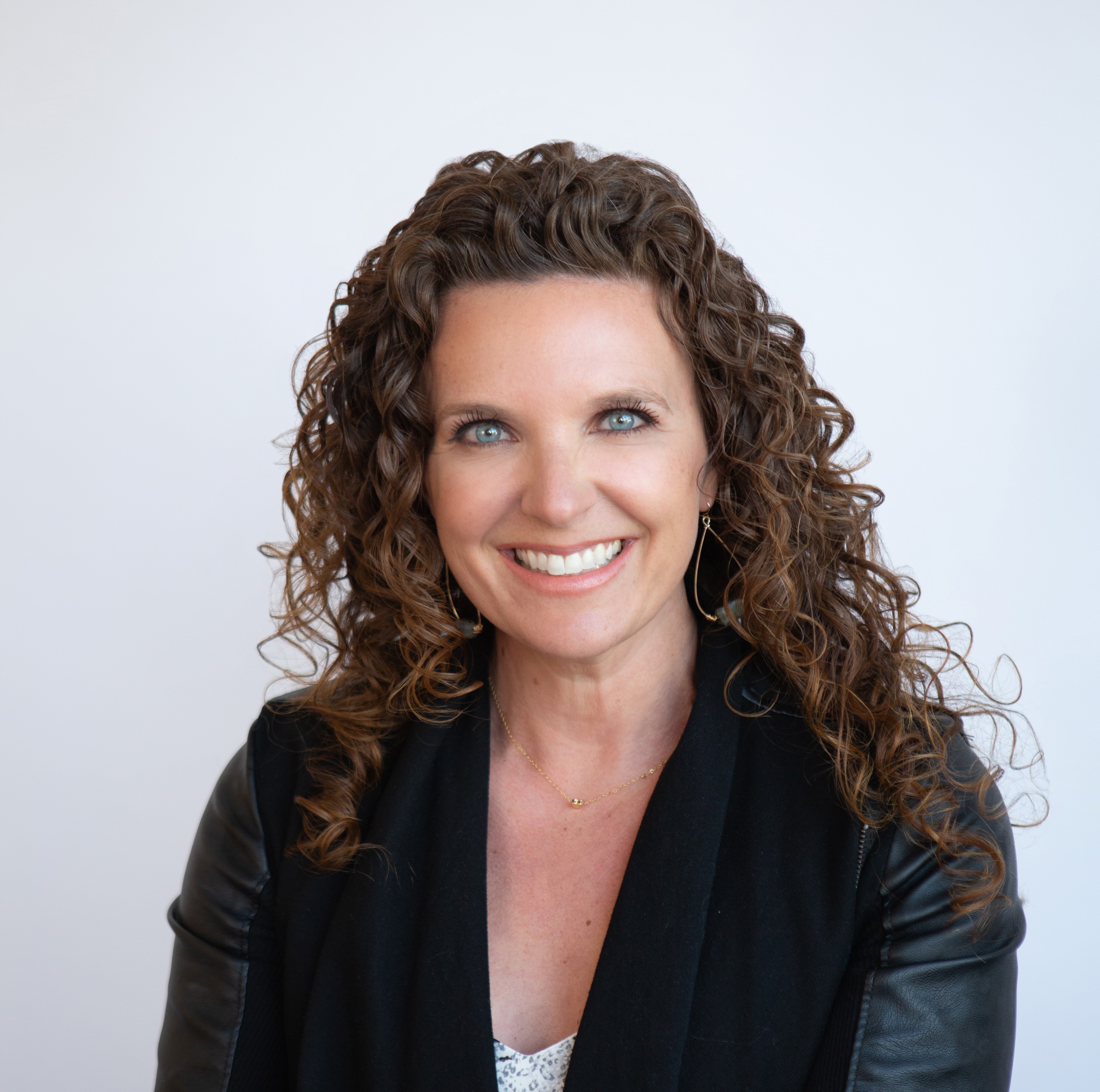 Woman with curly hair smiling, wearing a black jacket, against a plain light background.