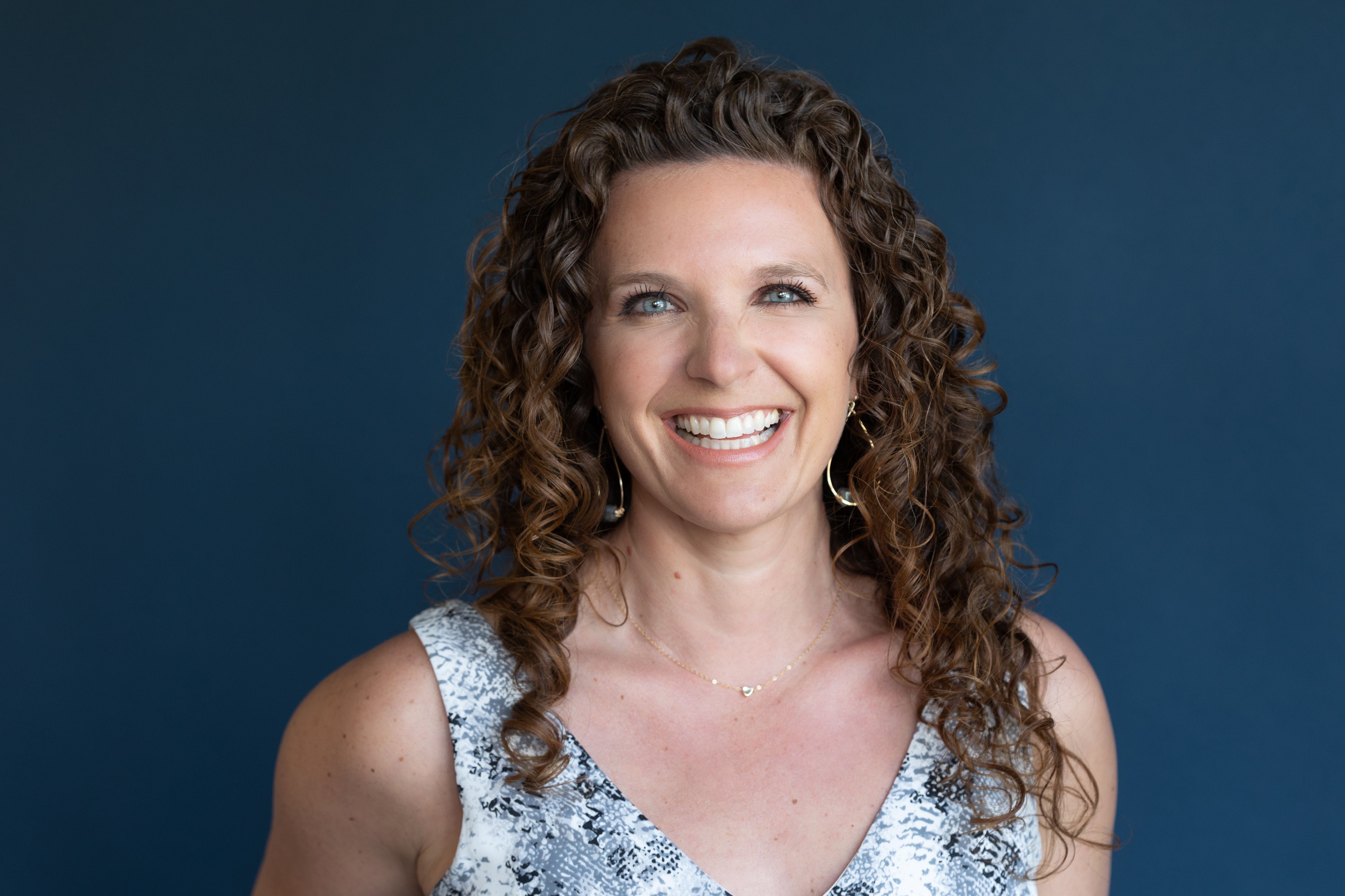 Woman with curly hair smiling, wearing a sleeveless top, against a dark blue background.