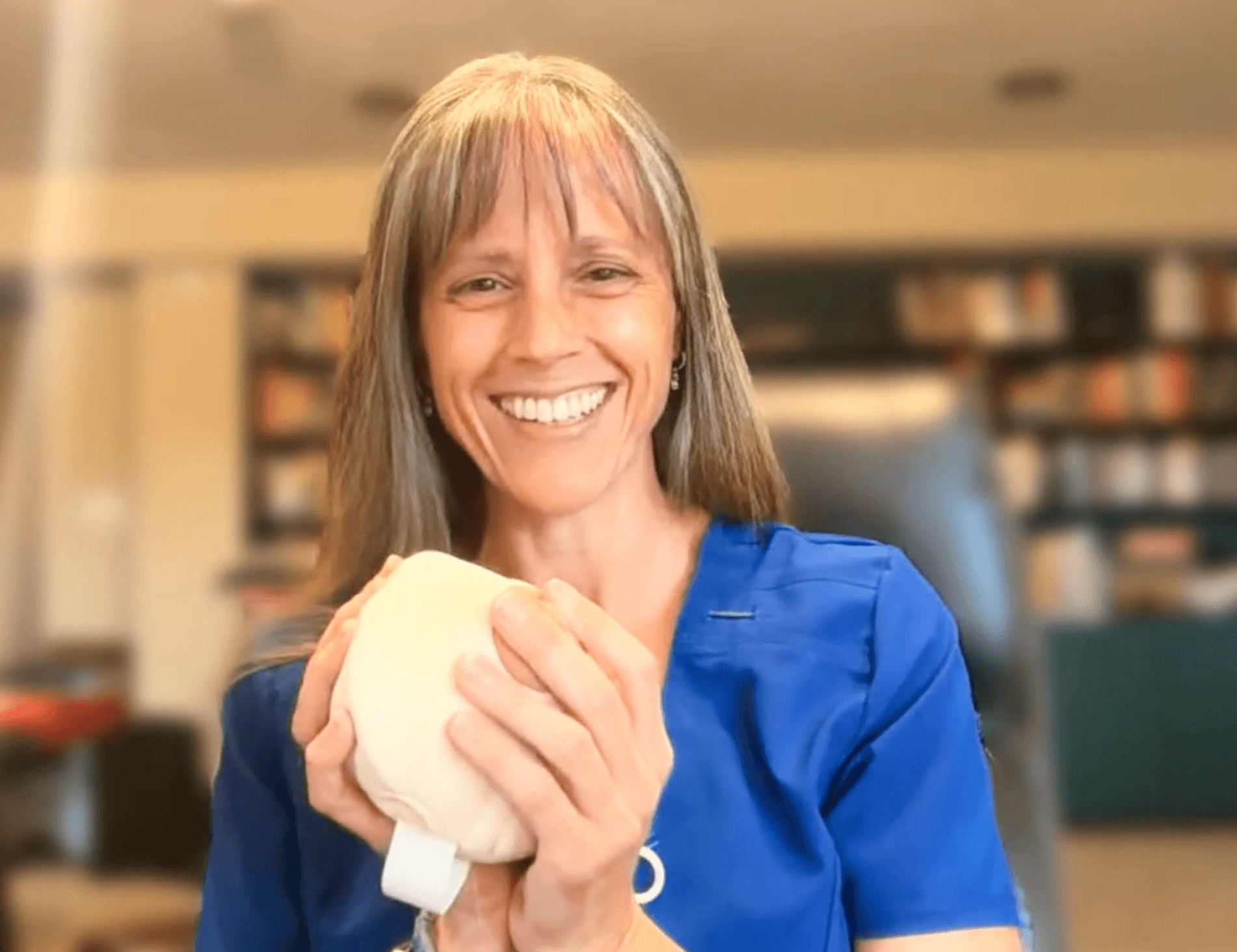 Smiling woman in blue scrubs holding a white object, in a room with bookshelves in the background.