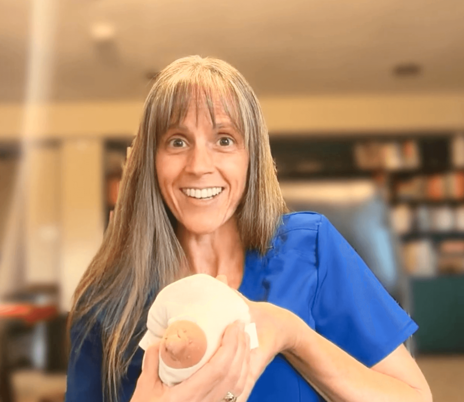 Woman with long hair smiling and holding a round object in a room with bookshelves in the background.
