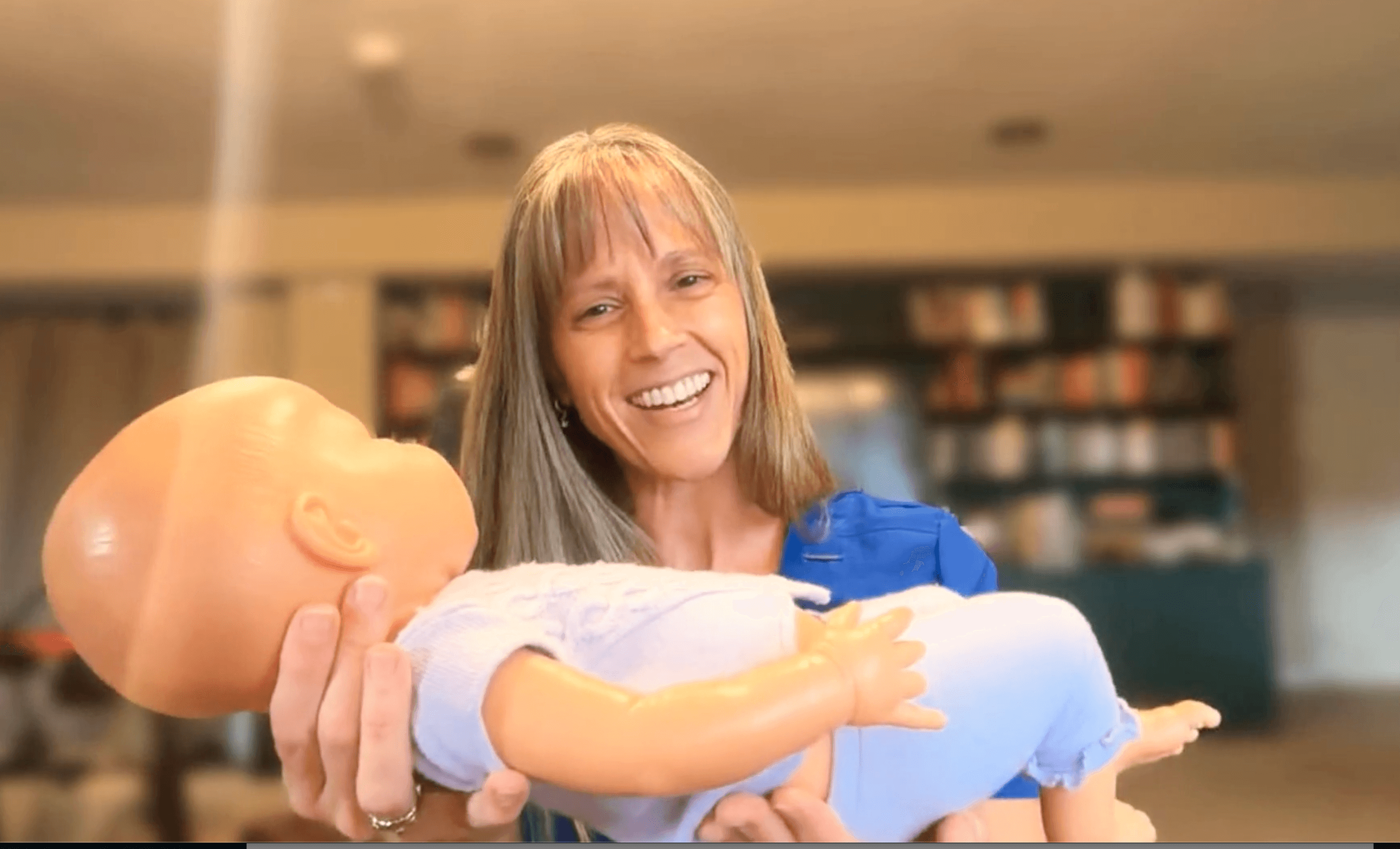 Woman smiling and holding a baby doll indoors, background with bookshelves, warm lighting.