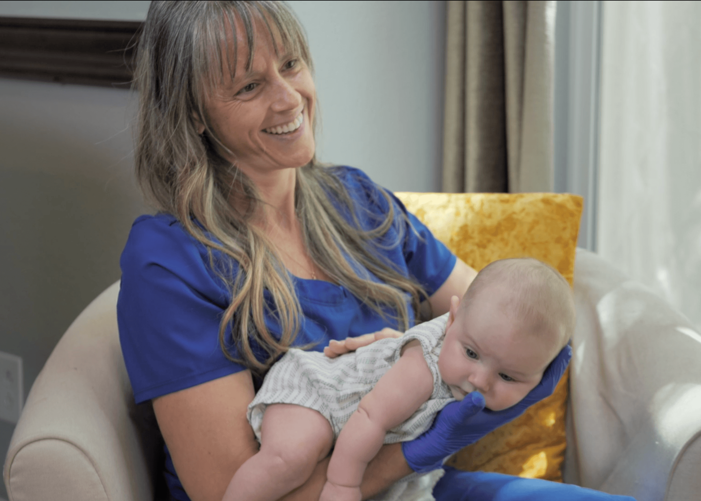 Woman holding a baby on her lap, smiling, in a room with a yellow pillow and curtains.