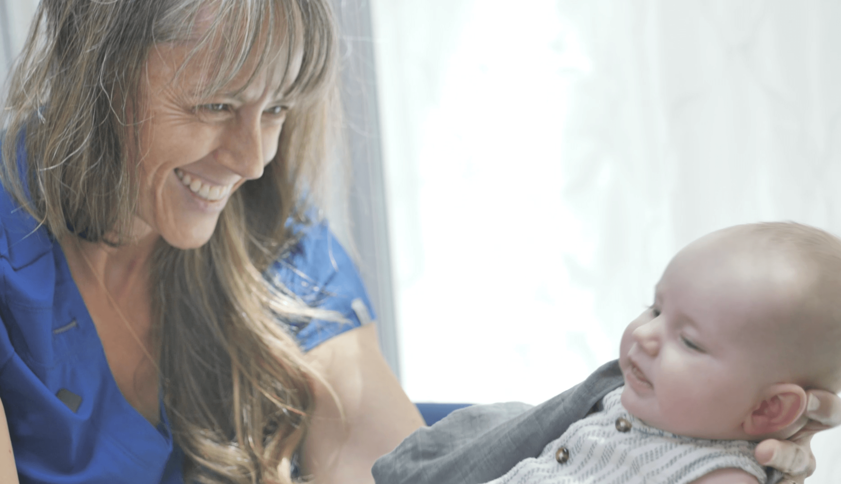 Woman smiling at a baby lying on a bed, bright background, woman wearing a blue shirt.
