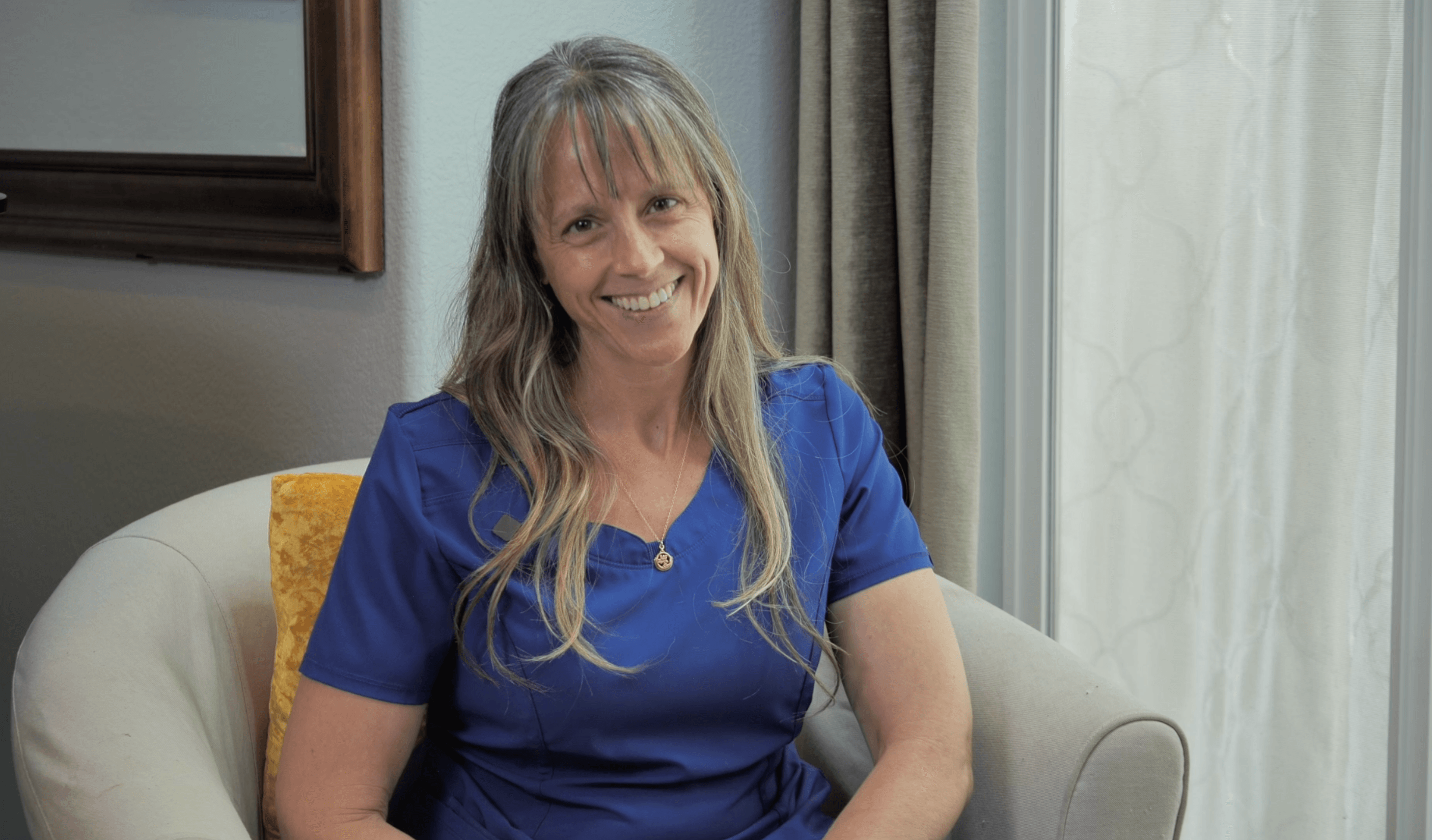 Smiling woman with long hair in blue scrubs sitting on a beige chair in a room with curtains and a mirror