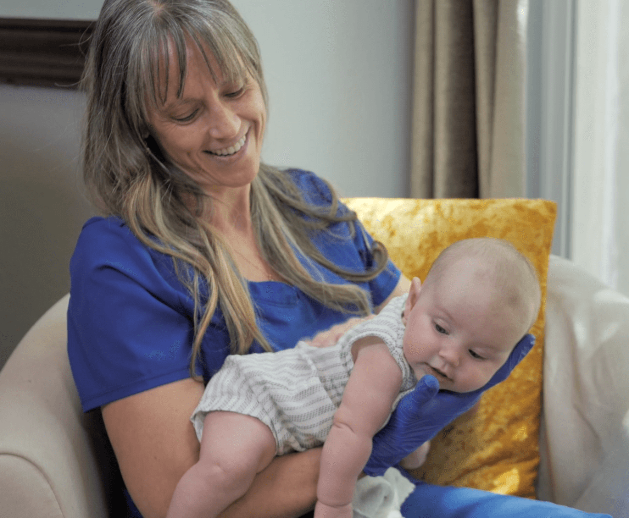 Woman holding a baby on her lap, smiling, in a cozy room with a yellow pillow and curtains.