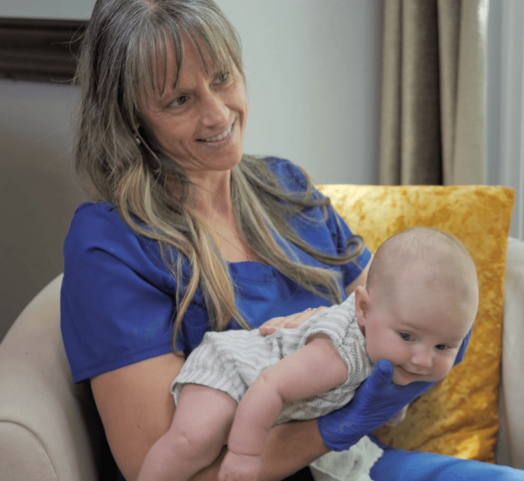 Woman holding a baby on her lap, sitting on a beige chair with a yellow pillow, smiling at the baby.