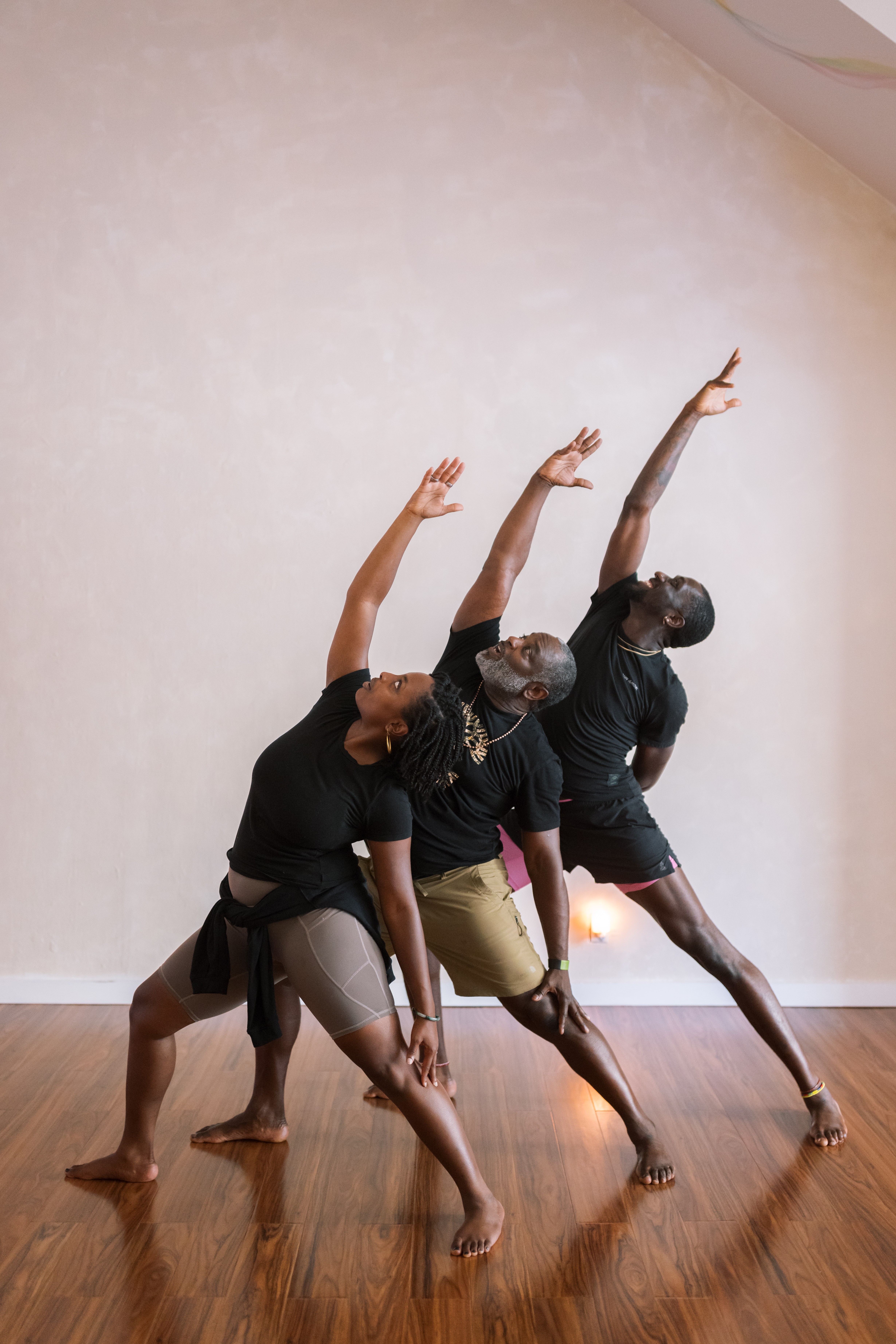 Three dancers in black outfits perform a synchronized pose with arms raised and bent, on a wooden floor against a plain wall.