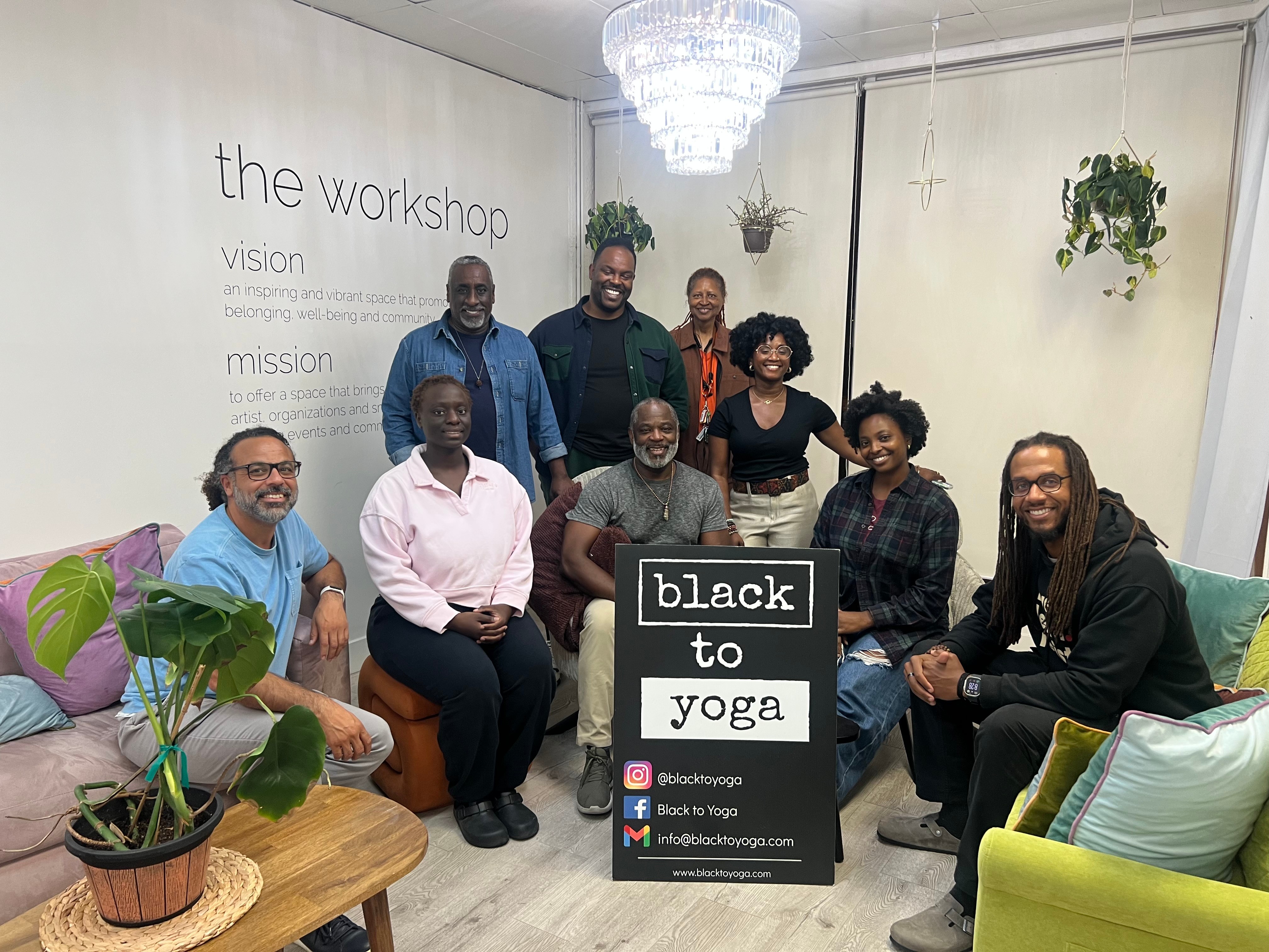 Group of nine people gathered in a room with a sign reading 'black to yoga' in front, plants, and a chandelier.