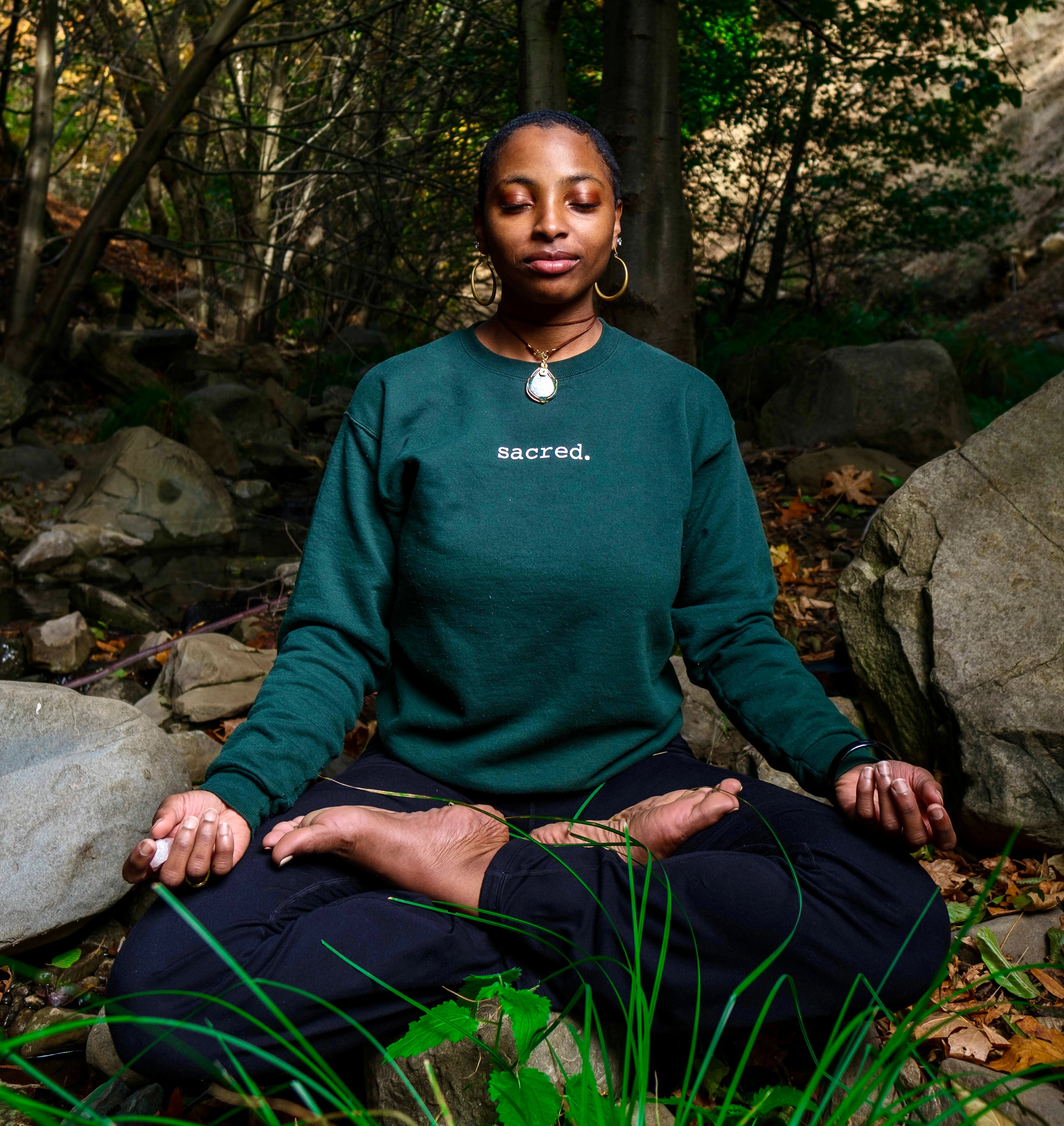 Woman meditating outdoors in a forest, sitting cross-legged with hands on knees, eyes closed, wearing a green sweatshirt with 'sacred' text.