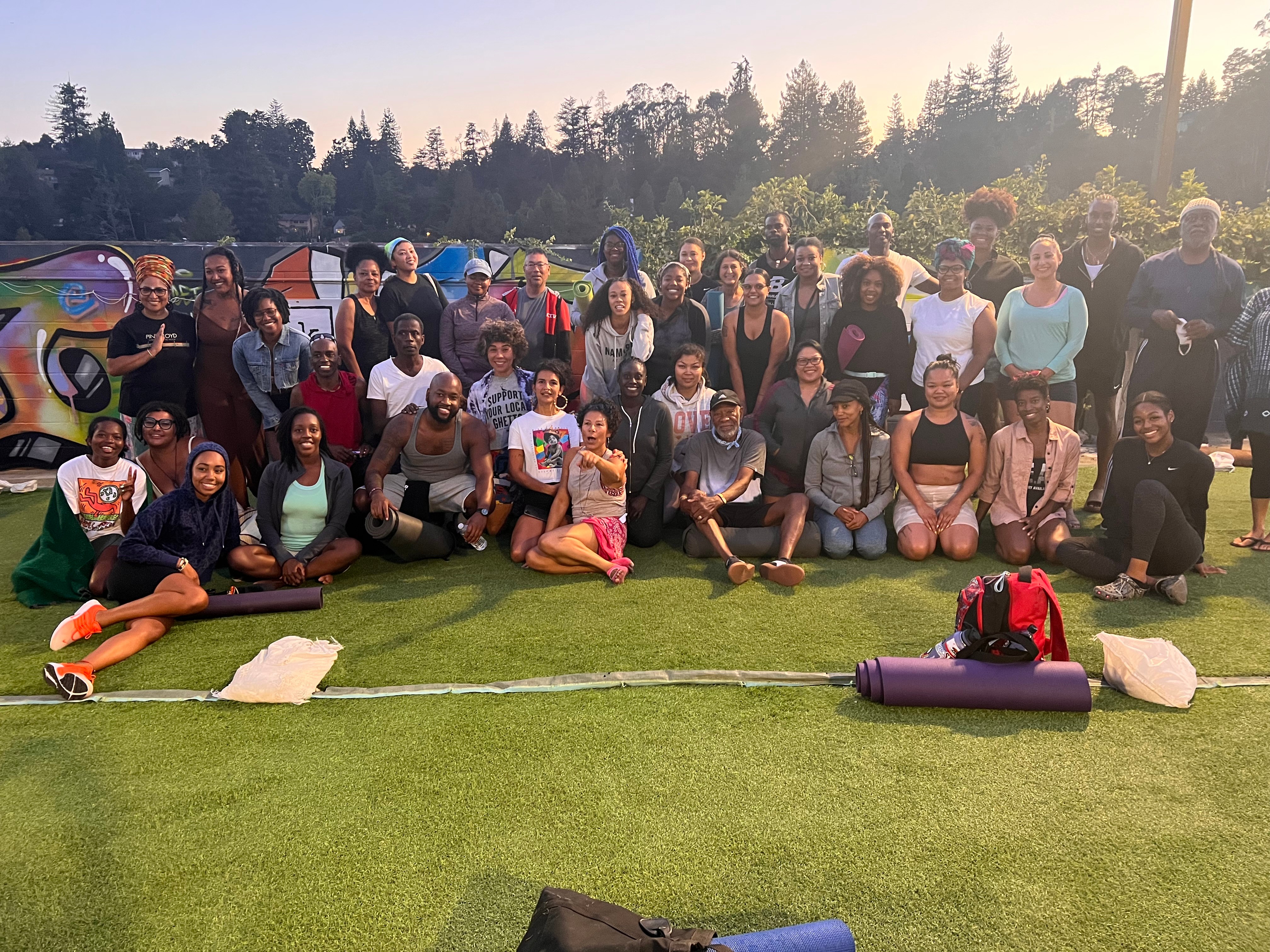 Group of people outdoors on grass, some sitting and some standing, with trees and tents in background.