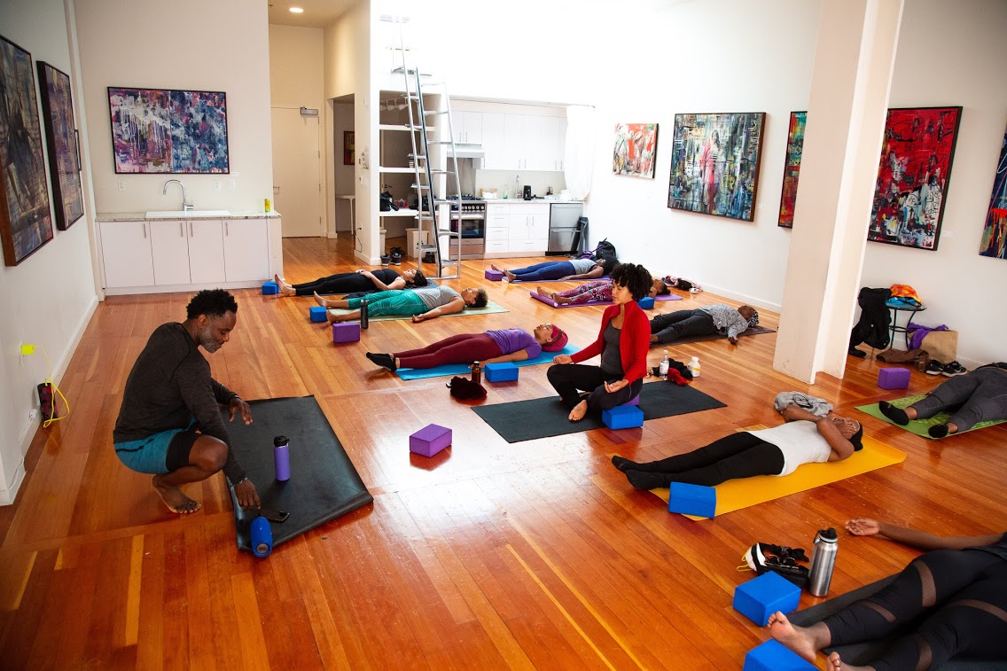 People participating in a yoga class in an art gallery with colorful paintings on white walls, wooden floor, and yoga mats.