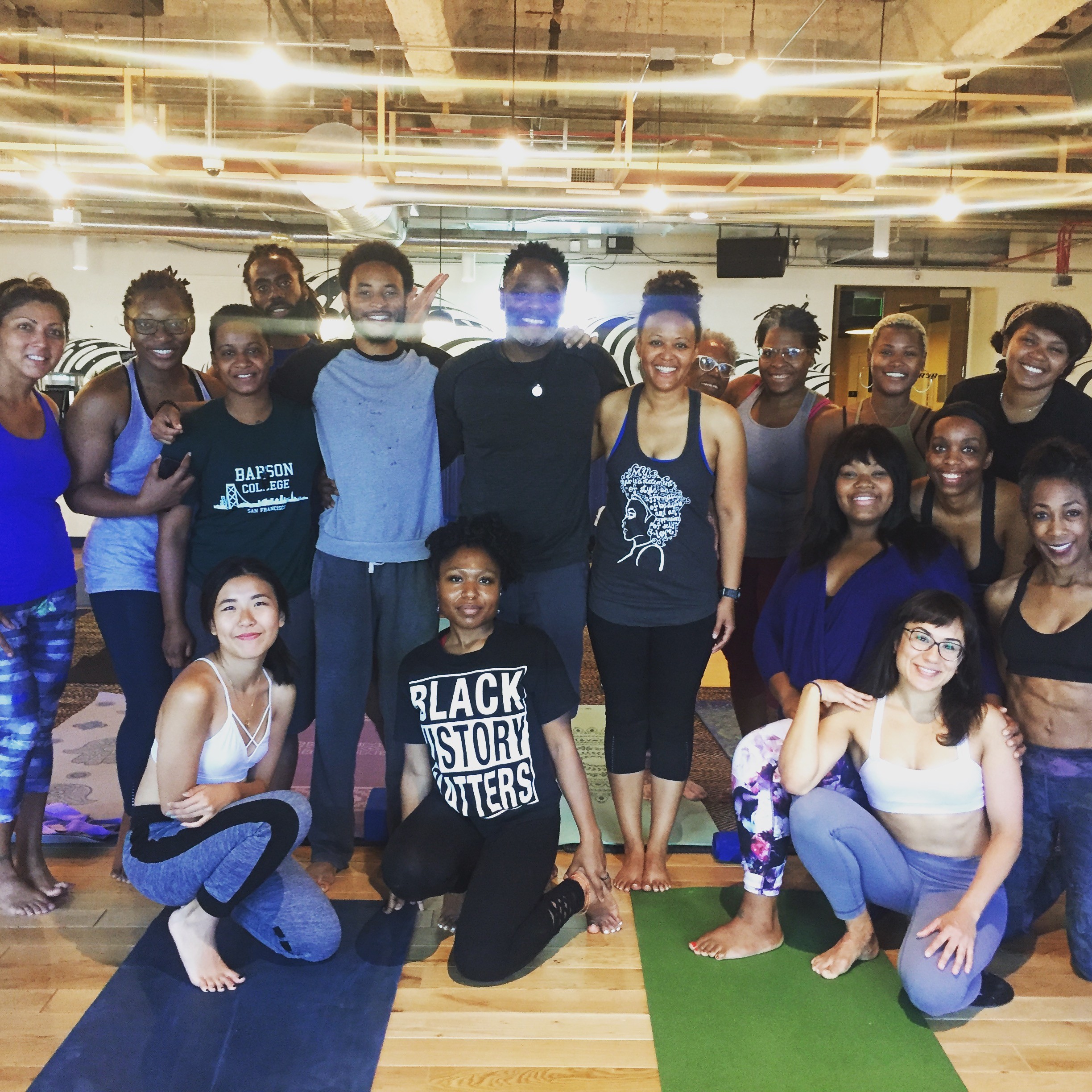 Group of people in a fitness studio, some standing and some sitting, smiling at the camera, with wooden floors and ceiling lights.