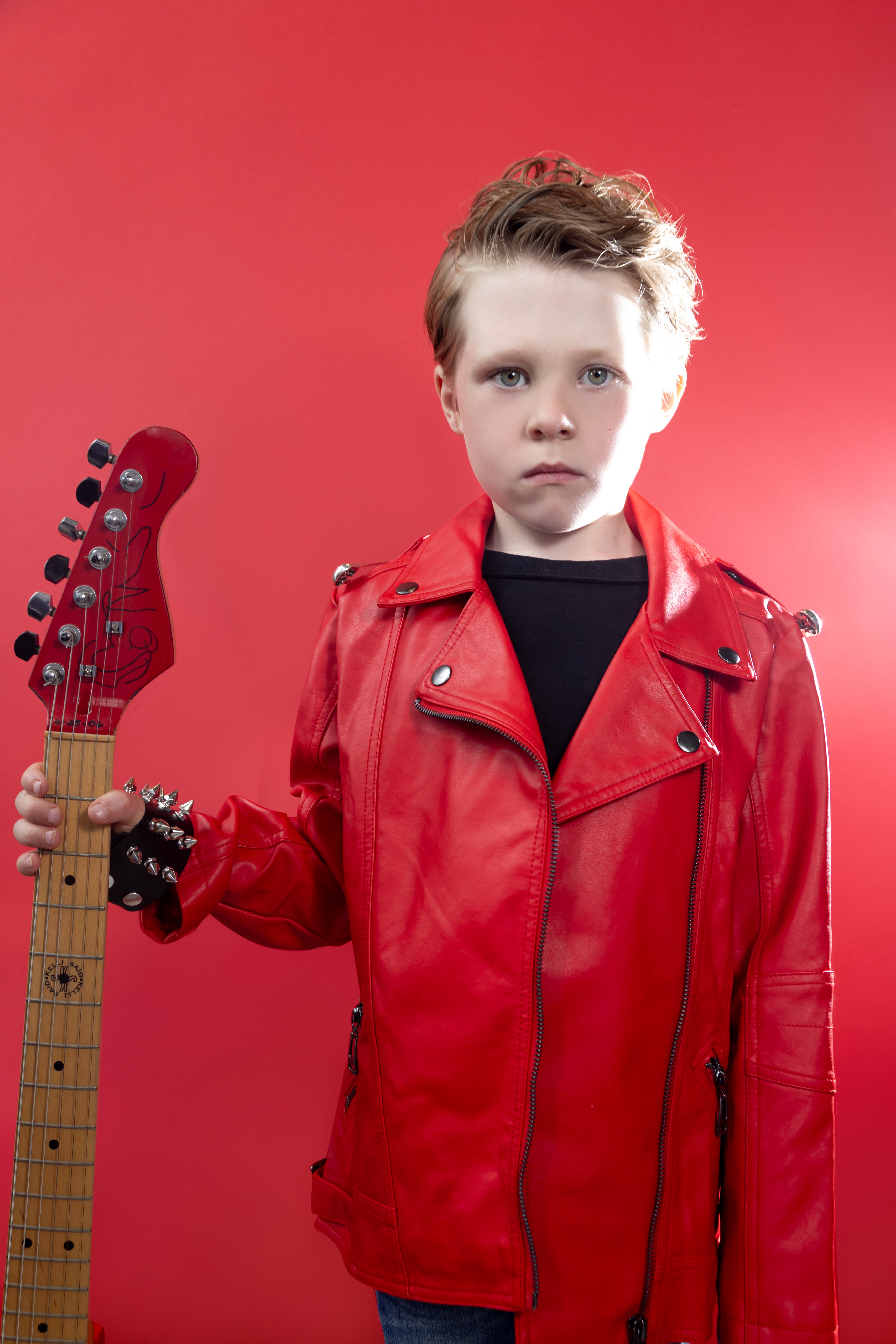 Young boy with a serious expression wearing a red leather jacket, holding a red electric guitar, against a red background.