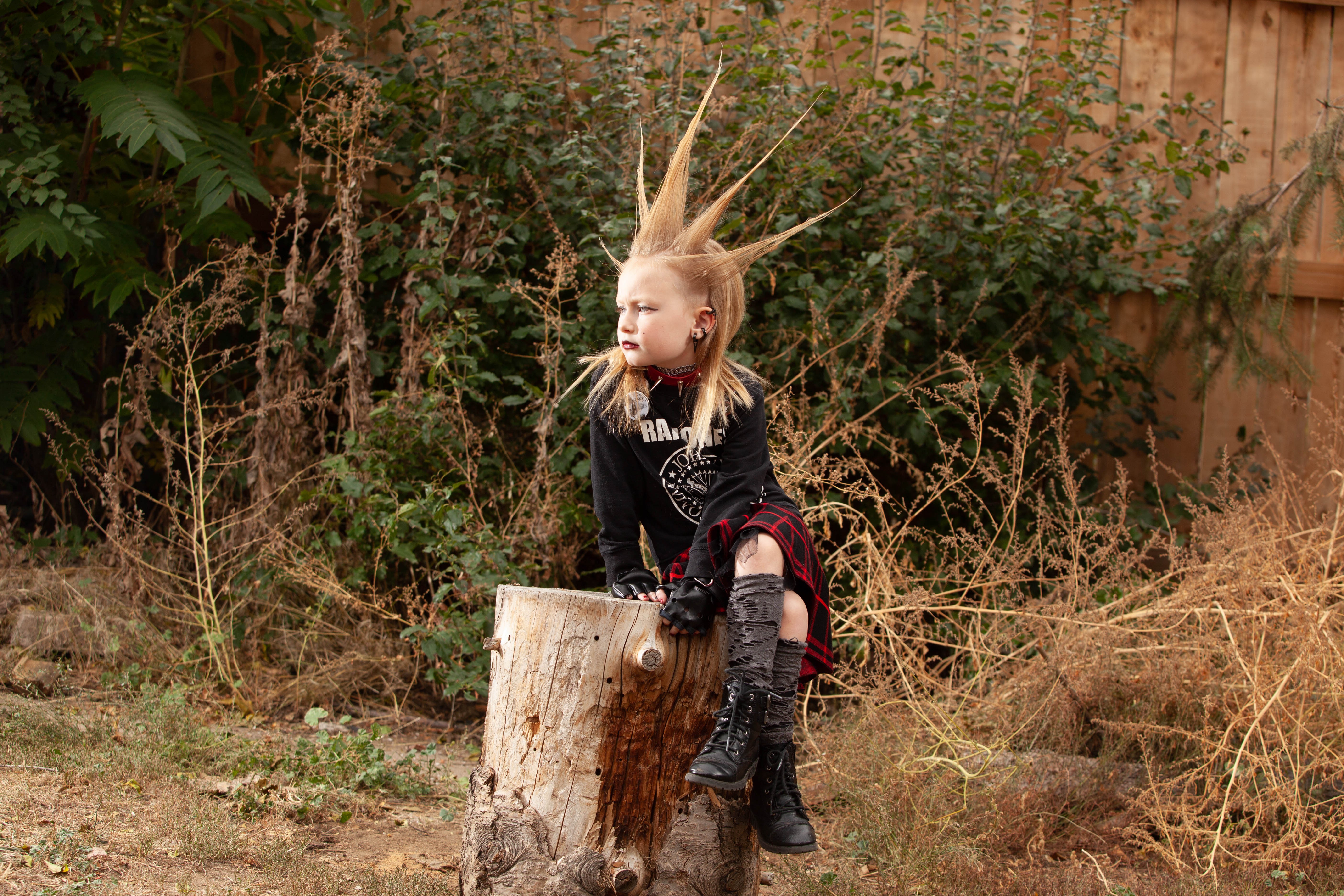 Child with spiked hair sitting on a tree stump outdoors, surrounded by dry grass and bushes.