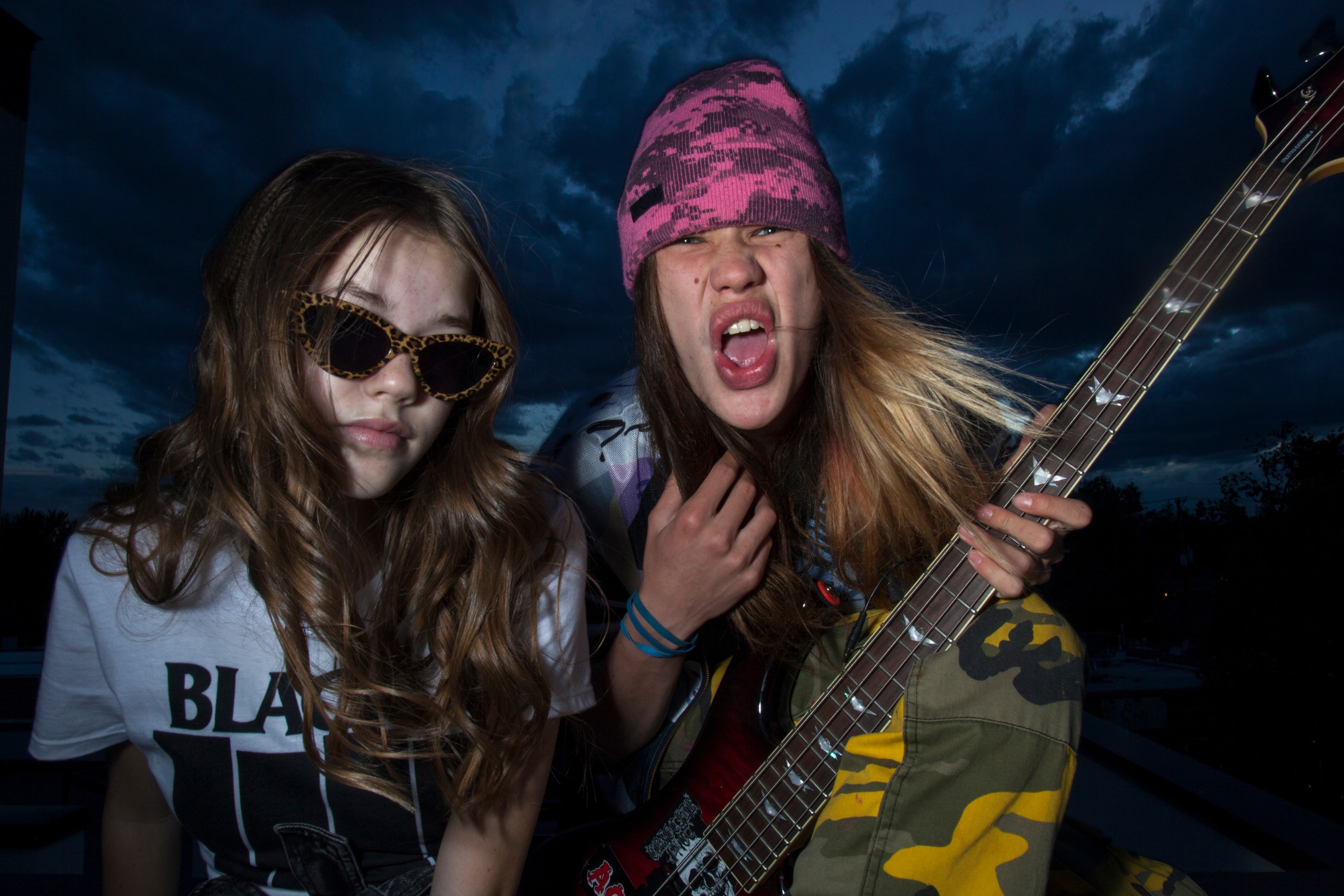 Two young women outdoors at night, one wearing sunglasses and the other with a pink bandana, holding a guitar, making expressive faces.