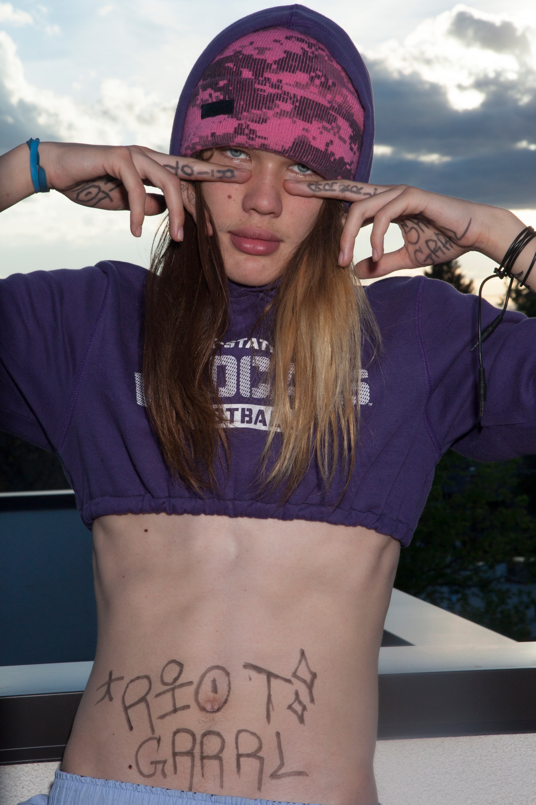 Young woman with a pink and purple cap, wearing a purple hoodie, covering her eyes with her hands, and writing on her stomach.