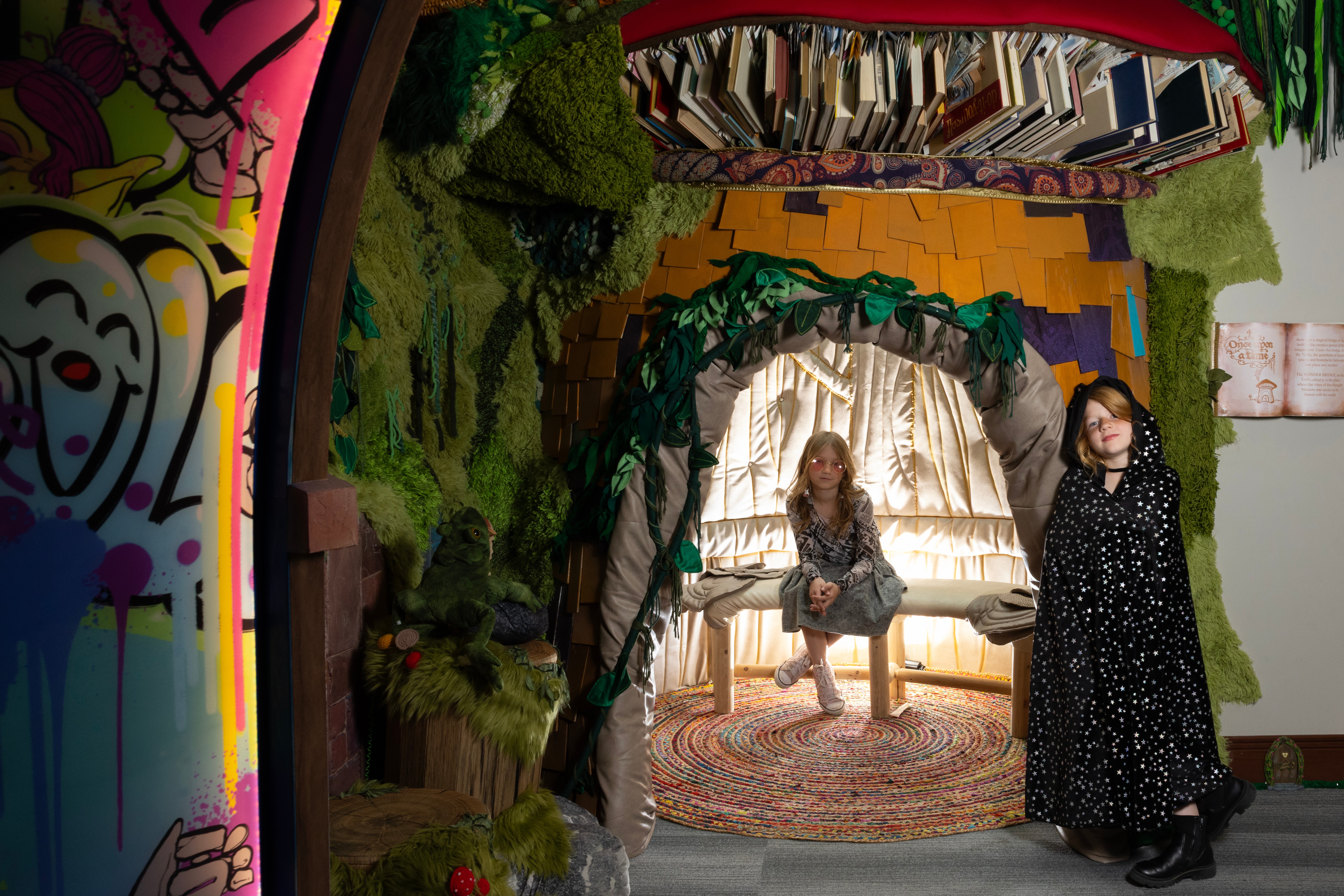 Two children on a stage with a forest backdrop, one sitting on a bench and the other standing, under a green arch.