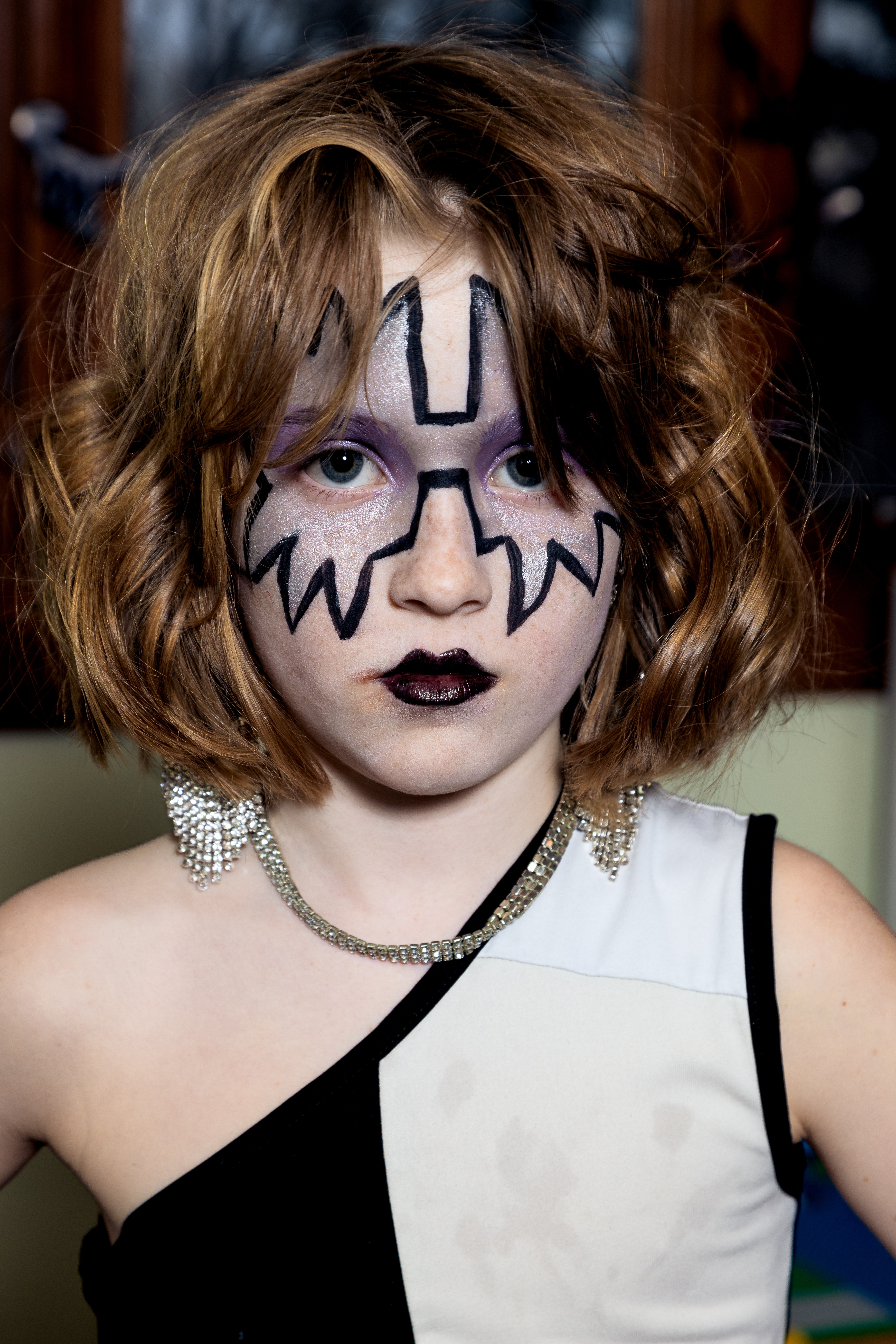 Person with face paint, dark lipstick, and curly hair, wearing a black and white outfit with jewelry.