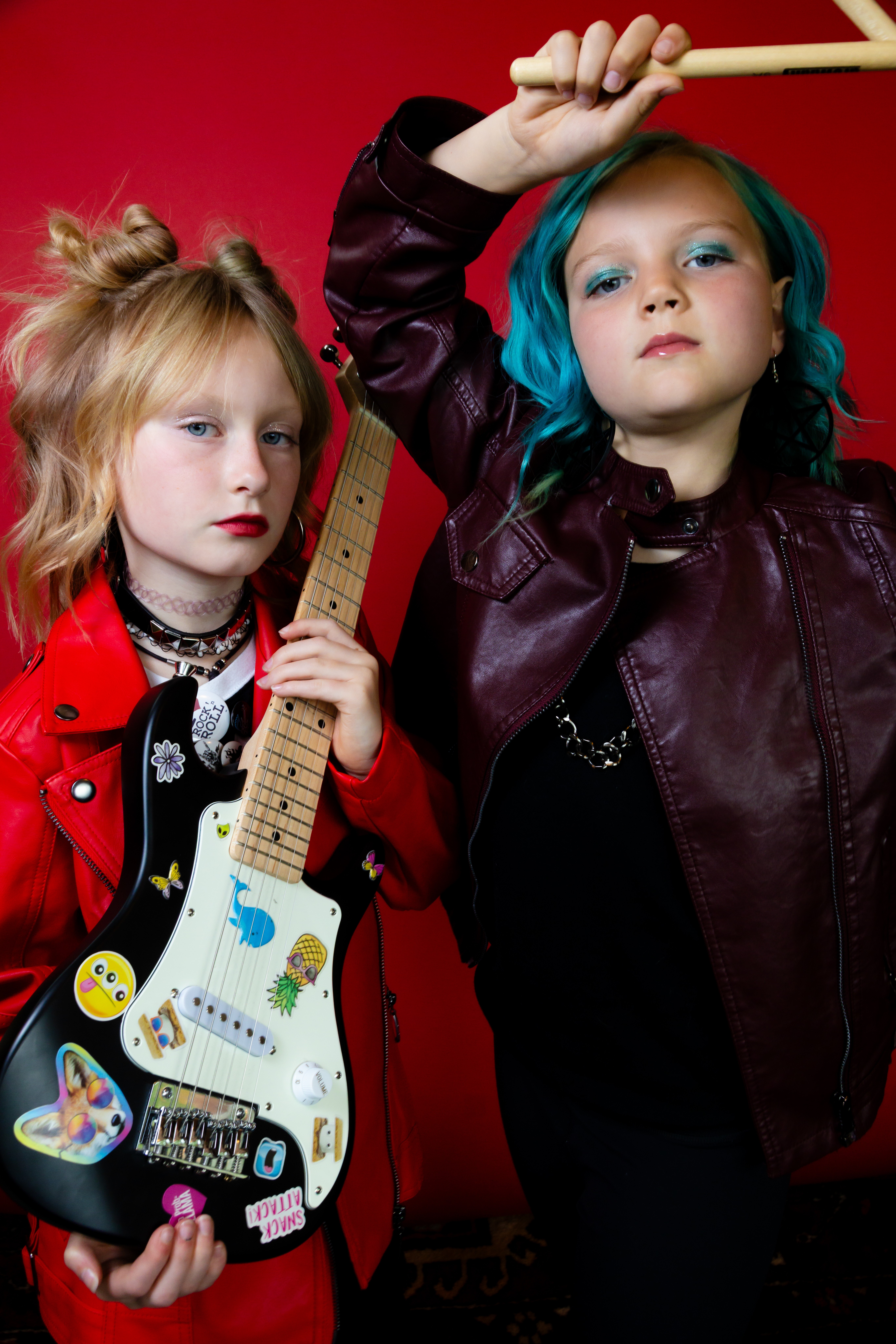 Two young girls with colorful hair and makeup, holding guitars and posing against a red background.